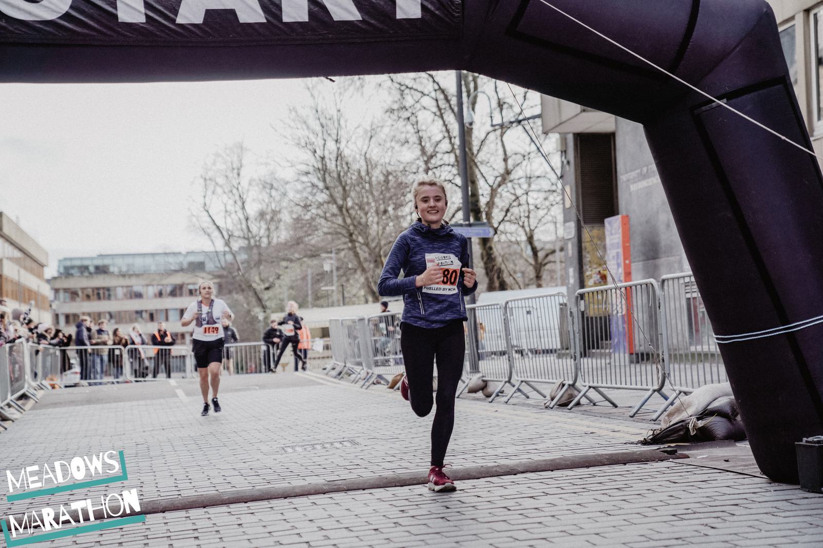 A runner with number 30 on their bib smiles as they cross the finish line at an outdoor marathon event. Other participants and spectators can be seen in the background. The event banner reads "Meadows Marathon.