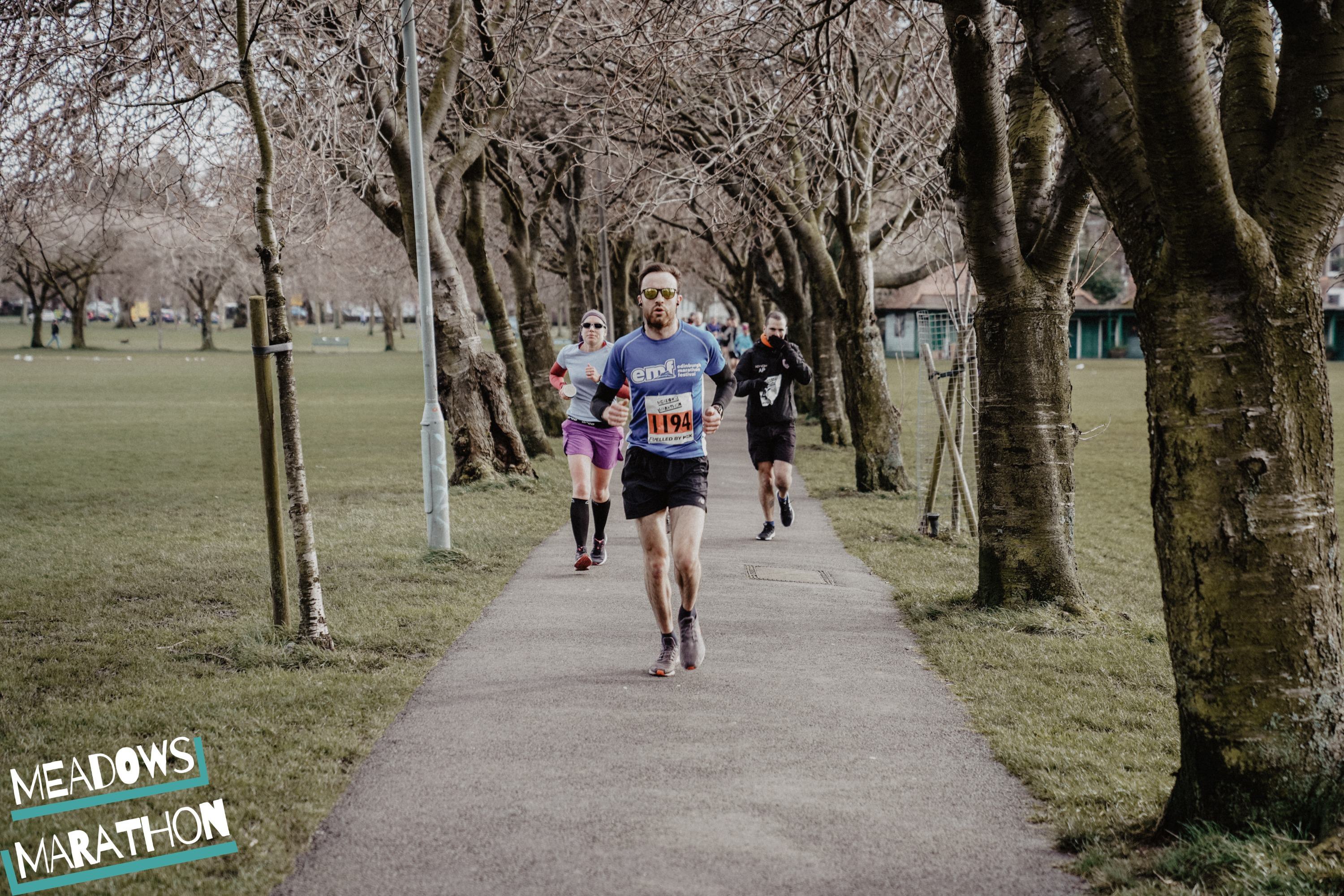 A group of individuals running on a paved path in a park, with leafless trees lining both sides. One runner, wearing a numbered bib, is leading while others follow behind. A "Meadows Marathon" sign is visible in the bottom left corner of the image.