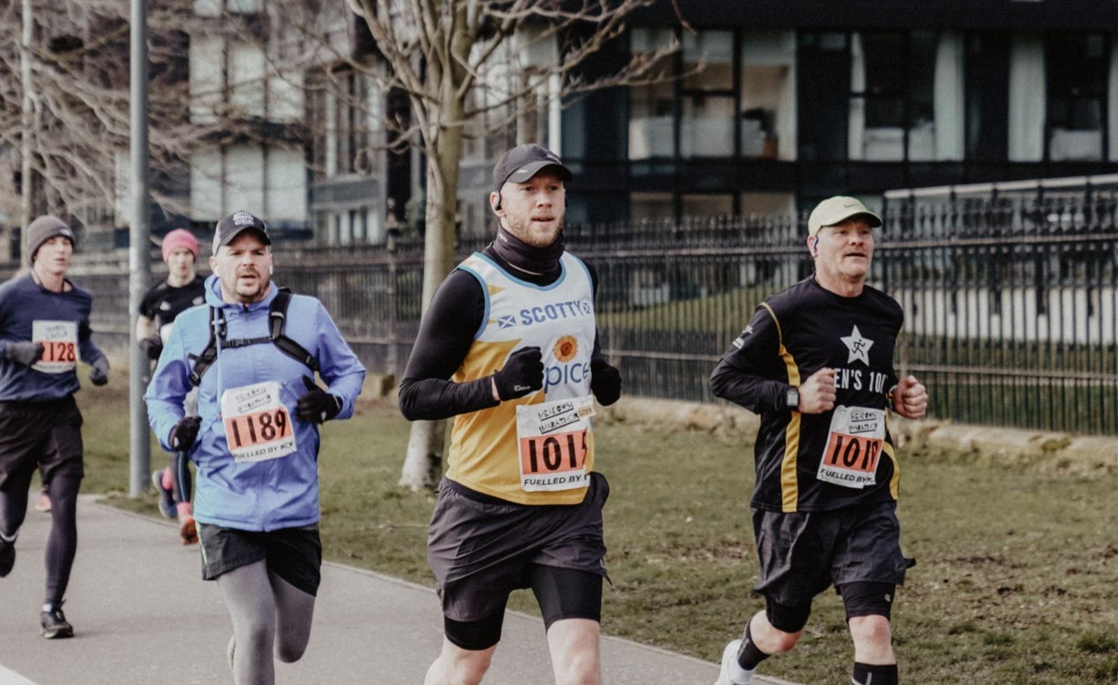 A group of runners dressed in athletic gear run on a concrete path during a marathon. They wear numbered bibs on their chests, and some are bundled up in hats and gloves. Trees and buildings line the route in the background, reflecting a cold weather setting.