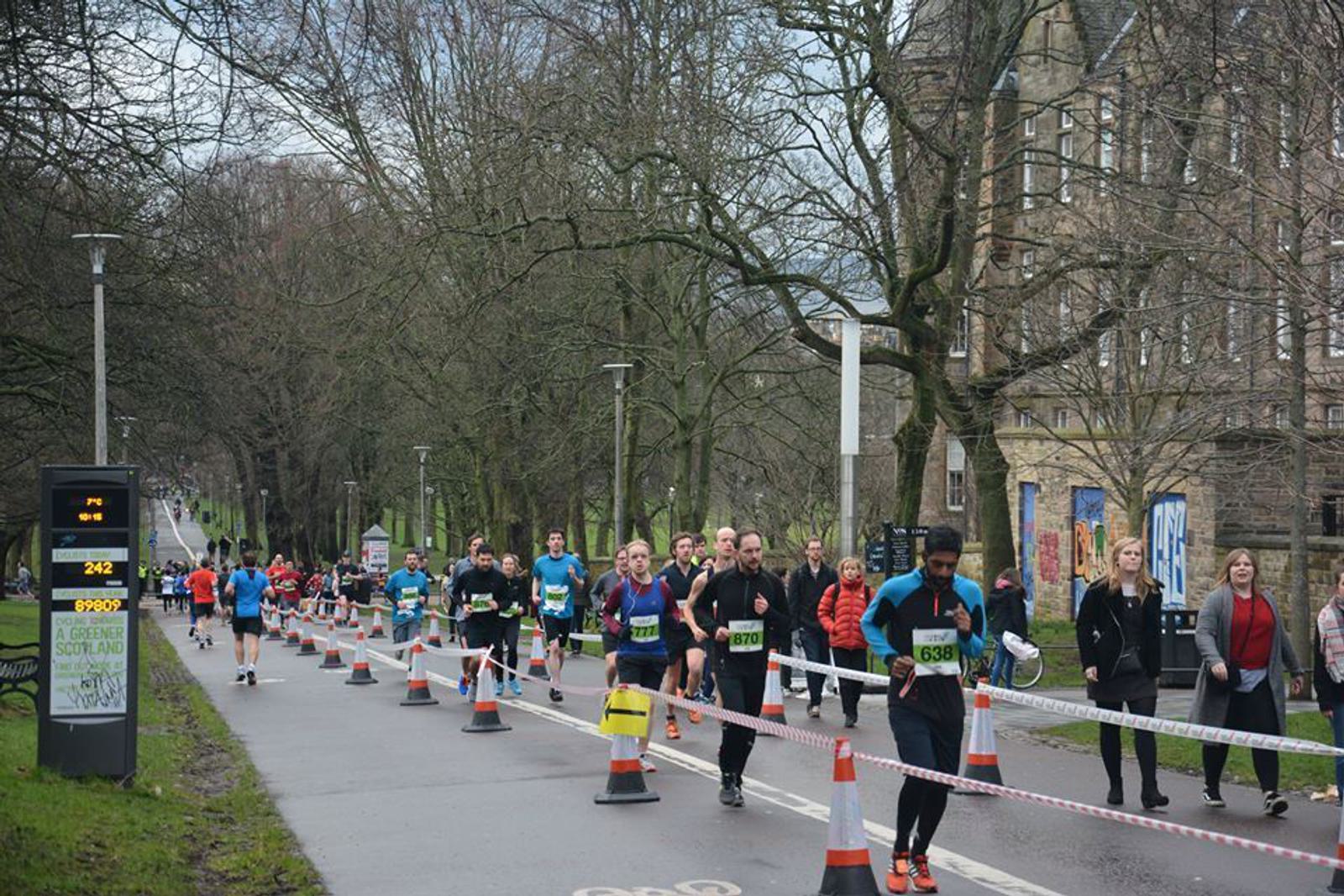 A group of runners in a race on a city street lined with trees. There are bystanders and spectators on the sidewalk. Several traffic cones and barriers are set up to mark the race route. Overcast sky and participants wearing numbers on their shirts.