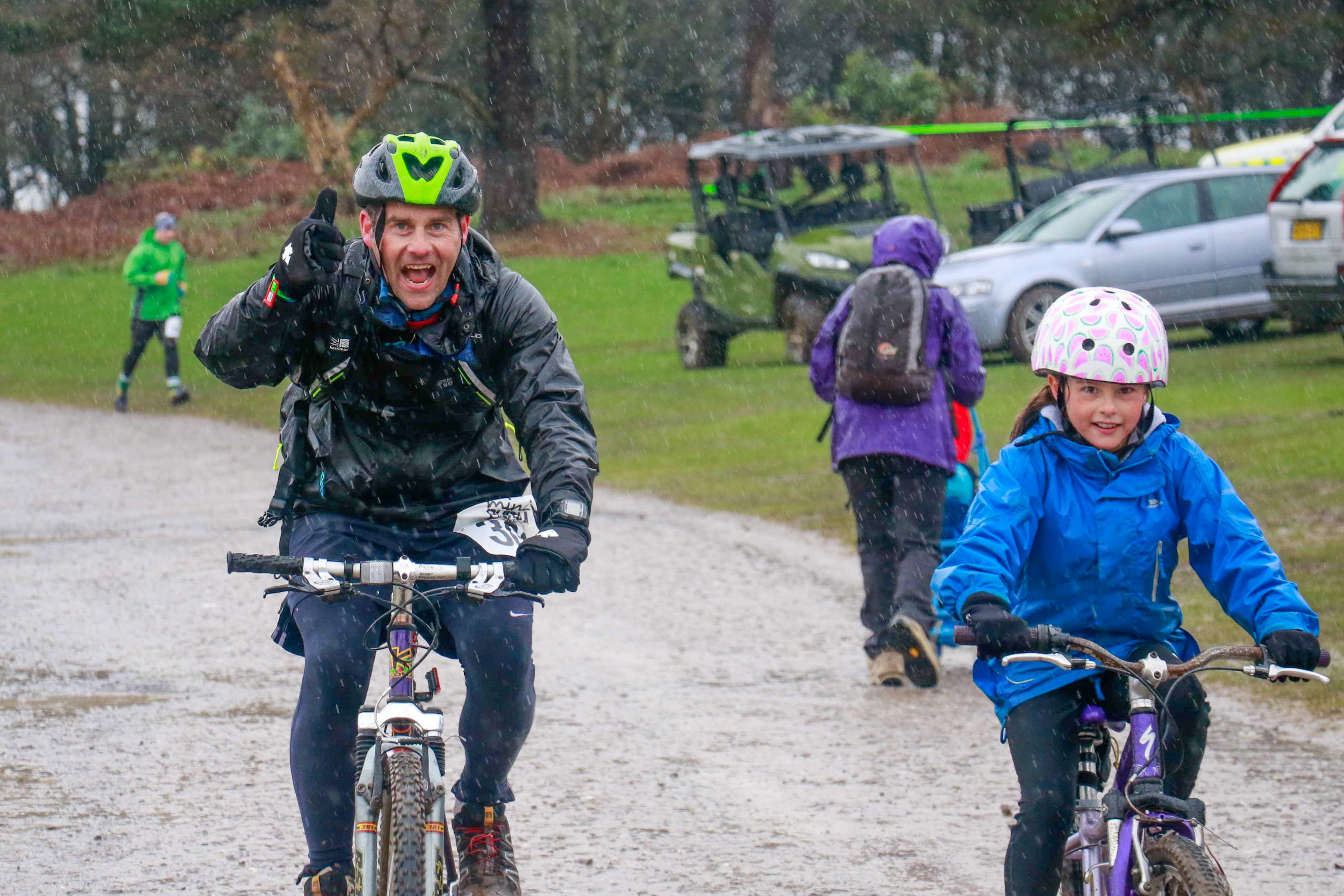 A smiling adult and a child, both wearing helmets and outdoor gear, ride bicycles on a rainy, muddy path. The adult gives a thumbs-up. Other people and parked vehicles are visible in the background, suggesting a group outdoor event or activity.