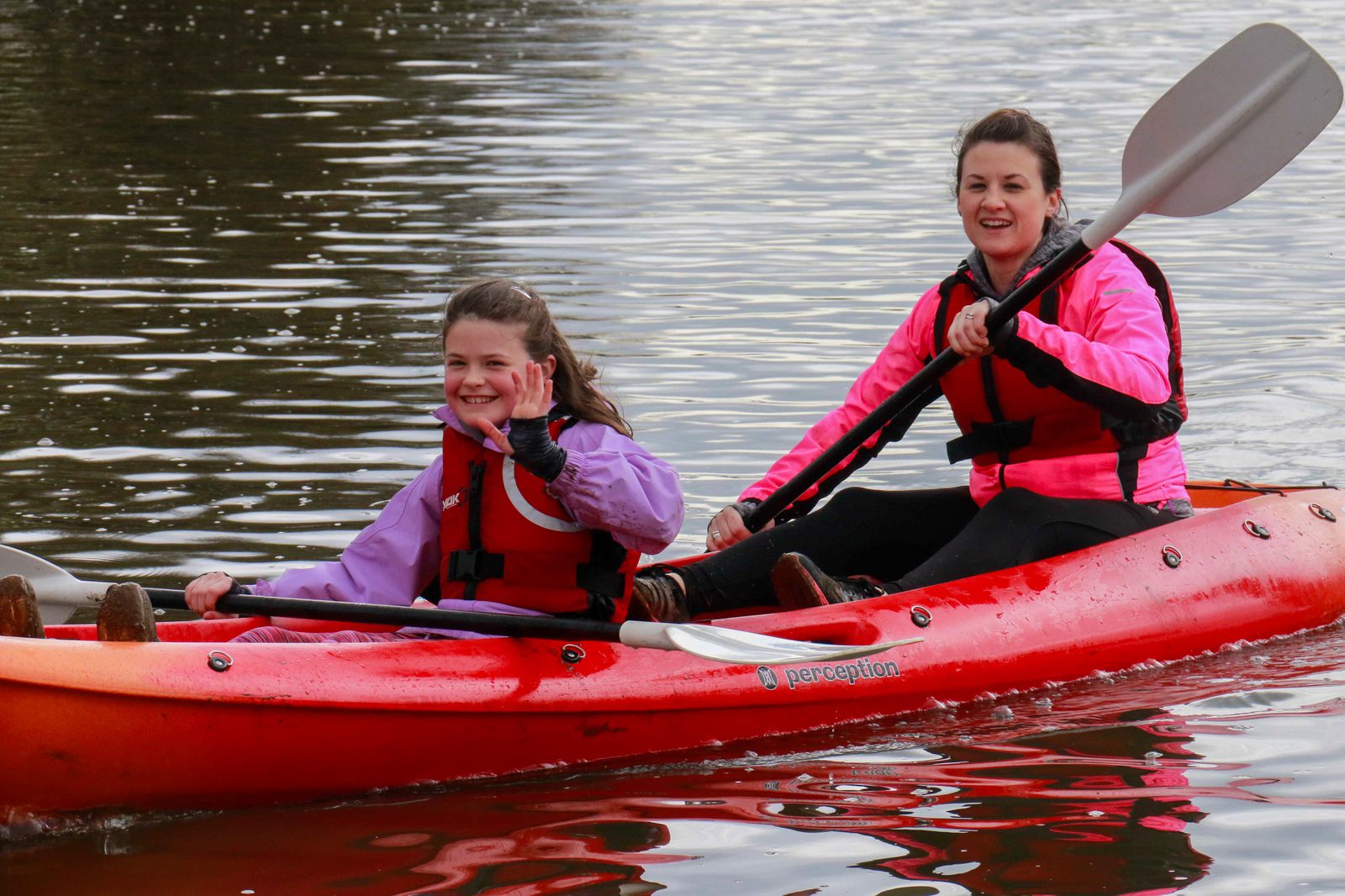 Two people are kayaking on calm water. Both are wearing life vests; the front person is waving and smiling, while the person in the back is paddling. They are in an orange kayak and appear to be enjoying the activity.