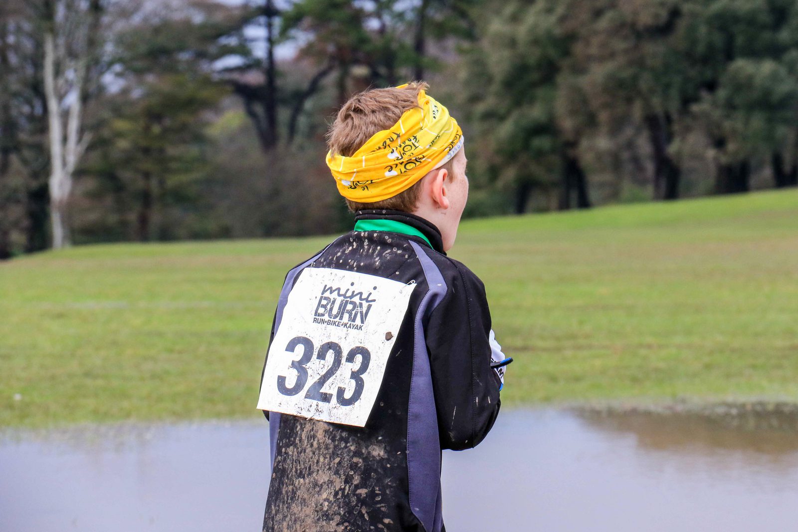 A person wearing a yellow headband and a race bib with the number 323 runs through a grassy area with patches of mud and water. Trees are visible in the background. The bib also shows the text "mini BURN.