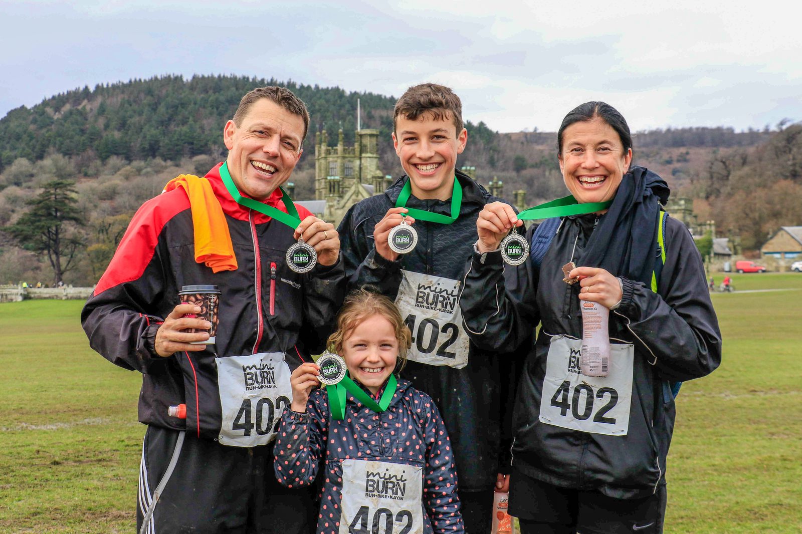 A group of four people, consisting of two adults, a teenager, and a young child, are standing outdoors, proudly displaying medals around their necks. They are all wearing race bibs with the number "402" and are smiling. The background shows trees and hills.