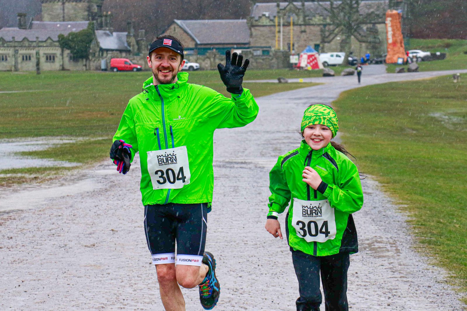 Two runners wearing neon green jackets and "304" bib numbers smile and wave as they run on a rainy day. The adult runner is on the left, wearing gloves and a cap, while the child runner is on the right. An old stone building and other runners are visible in the background.