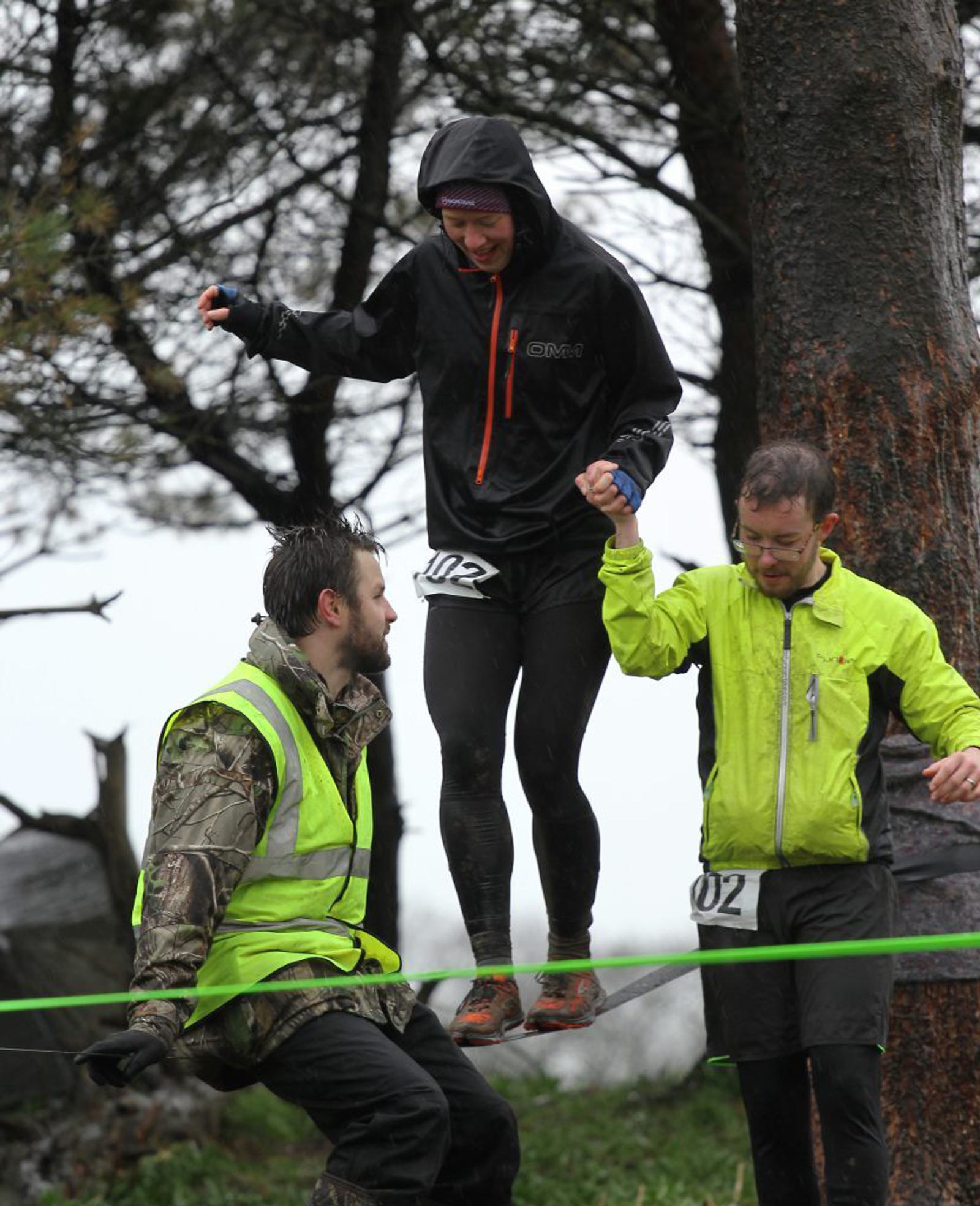 Three people participate in an outdoor race. One person in a black rain jacket and a numbered bib is balancing on a rope while being assisted by two others, one in a fluorescent yellow jacket and the other in camouflage gear. Trees and a wet environment are visible.
