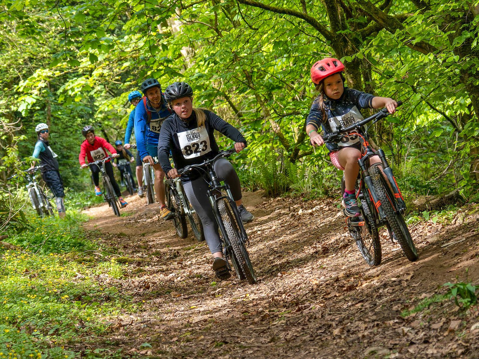 A group of cyclists, including children and adults, ride mountain bikes along a forest trail. The riders wear helmets and numbered race bibs, indicating participation in a biking event. The path is surrounded by lush greenery and trees.