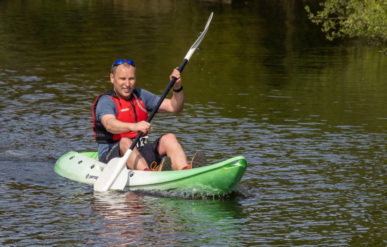 A person wearing a red life jacket and sunglasses is kayaking on a calm body of water. The individual is paddling a green kayak, creating small splashes as the paddle moves through the water. Trees and greenery are visible in the background.