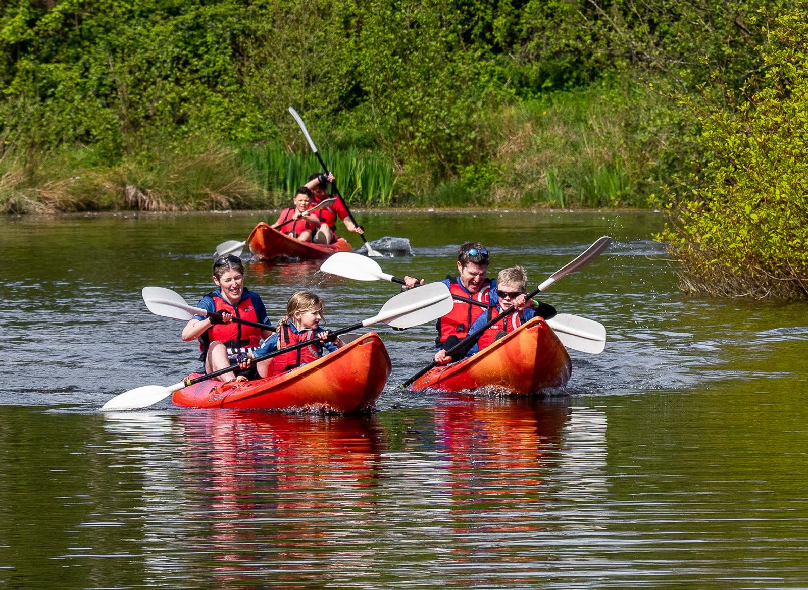 Four children in red life jackets paddle in pairs in red kayaks on a calm lake surrounded by green foliage. They seem to be enjoying a sunny day outdoors as they maneuver their kayaks through the water.
