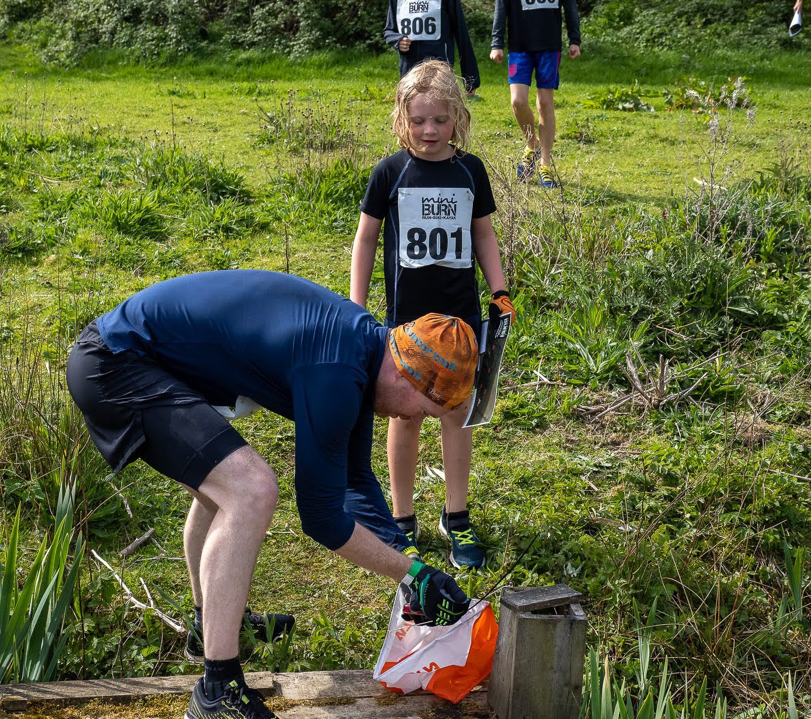 A man in athletic wear and an orange hat, bends over to check a checkpoint marker during an outdoor orienteering race. Nearby, a young girl with a race bib number "801" watches. Other participants are visible in the background amidst green foliage.