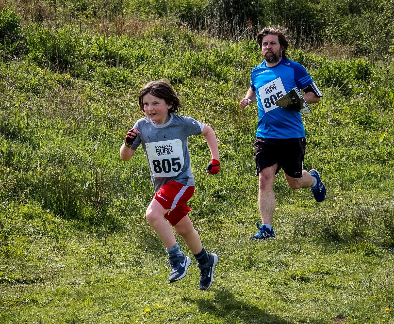 A boy wearing a grey shirt and red shorts, number 805, runs energetically on a grassy trail during a race. Behind him, an adult man in a blue shirt and black shorts, number 806, also runs on the same path. Both look determined, with lush greenery in the background.