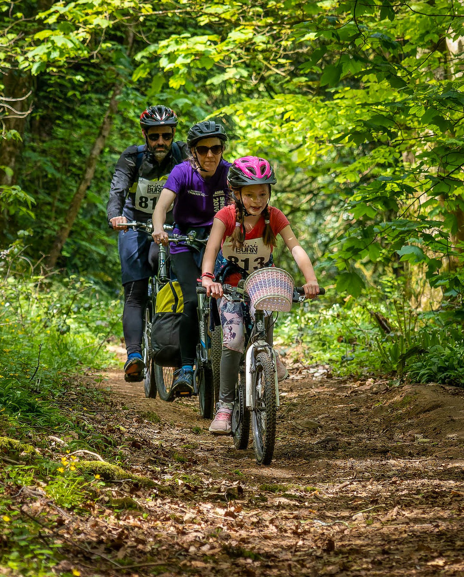 A young girl leads the way on a white bicycle, followed closely by two adults, all wearing helmets, riding along a narrow forest path surrounded by lush green foliage. The riders appear focused as they navigate the trail on a sunny day.