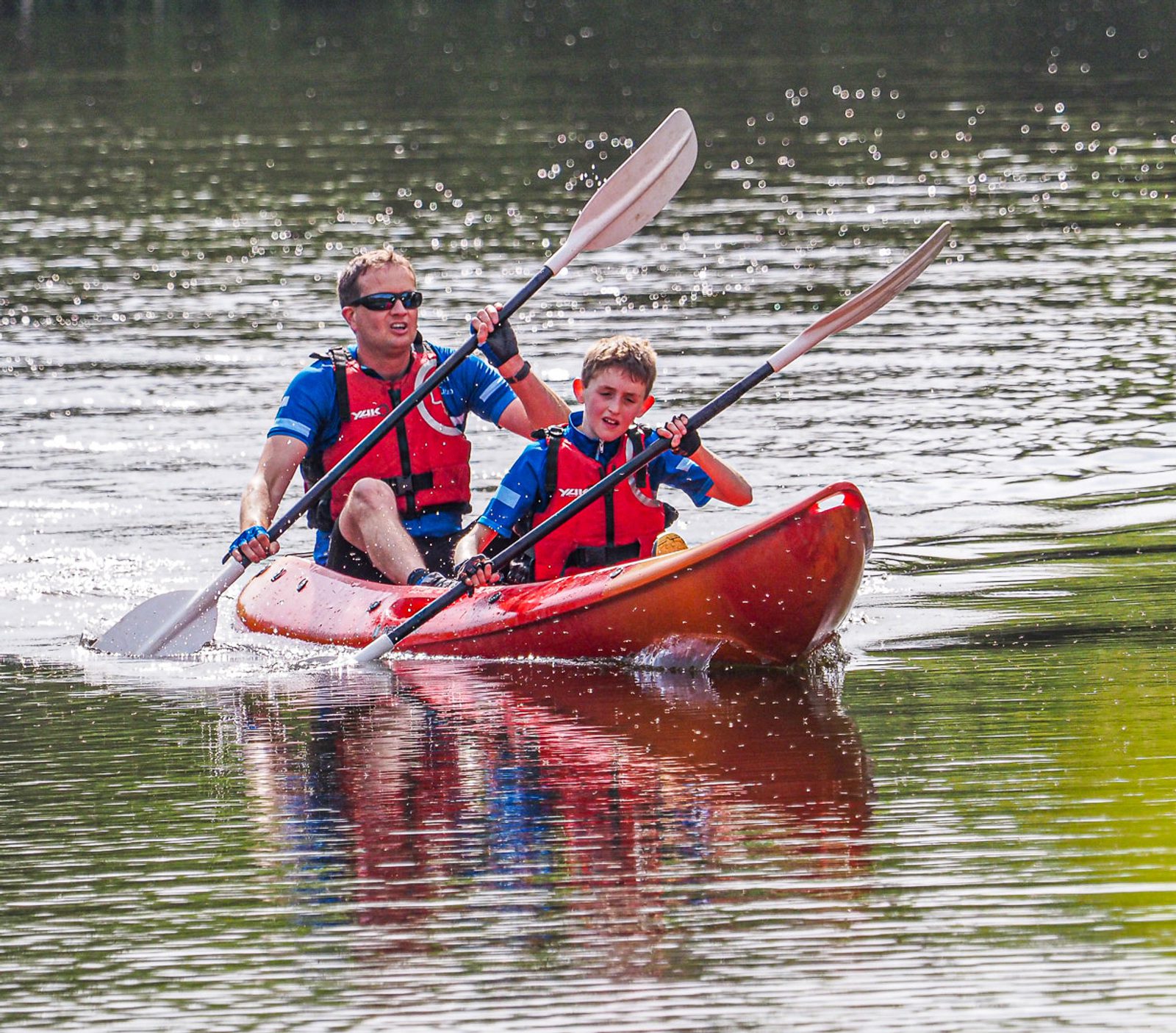 An adult and a child are kayaking on a calm body of water. Both are wearing blue shirts and red life jackets while paddling in unison. The background features reflective ripples, indicating a sunny day. The adult is seated at the back, guiding the child in front.