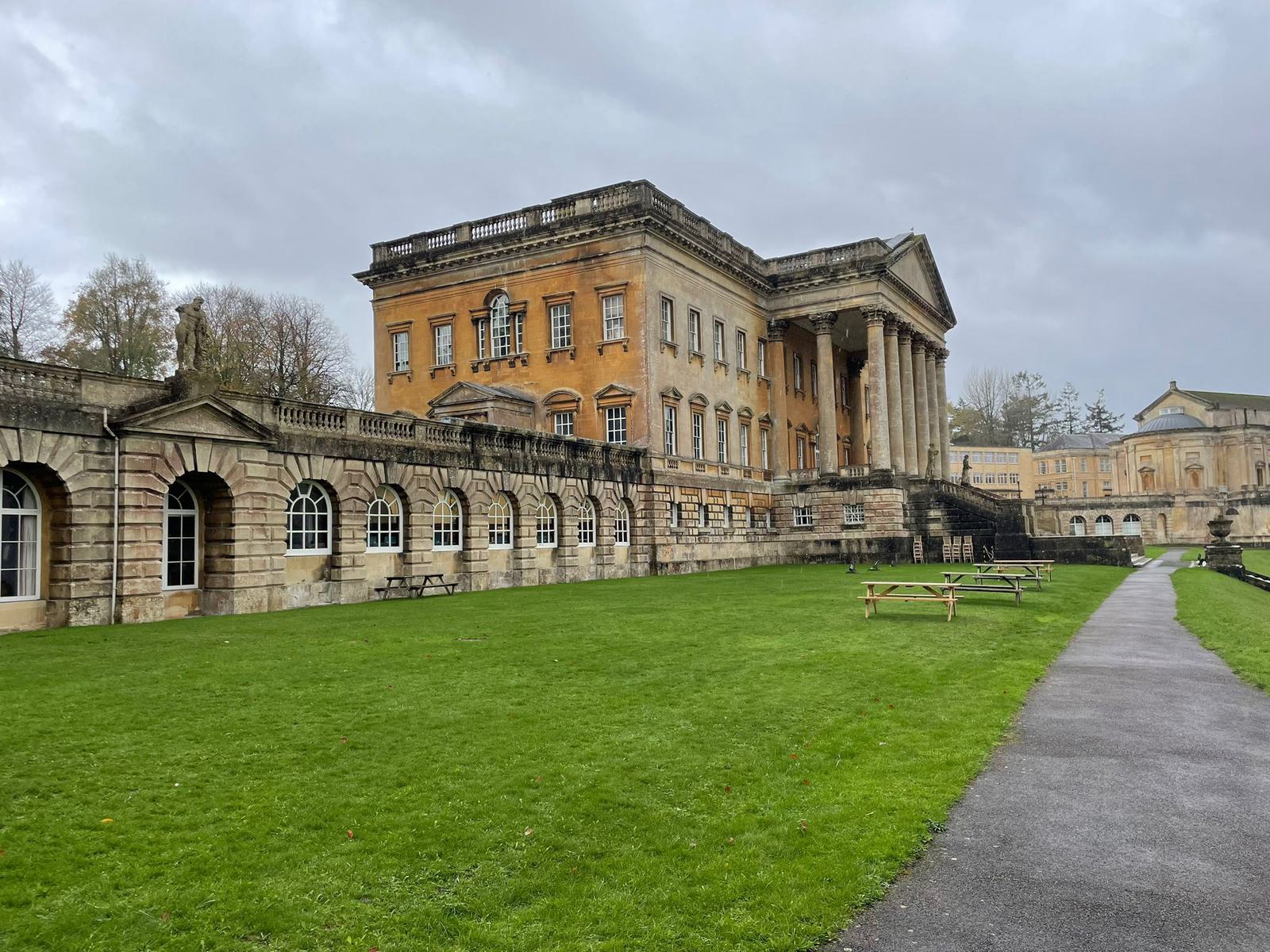 A large, historic building with classical architecture featuring columns and intricate stonework. The building has arched windows on the lower level. A neatly mowed green lawn and a paved path lead up to the entrance. Overcast sky in the background.