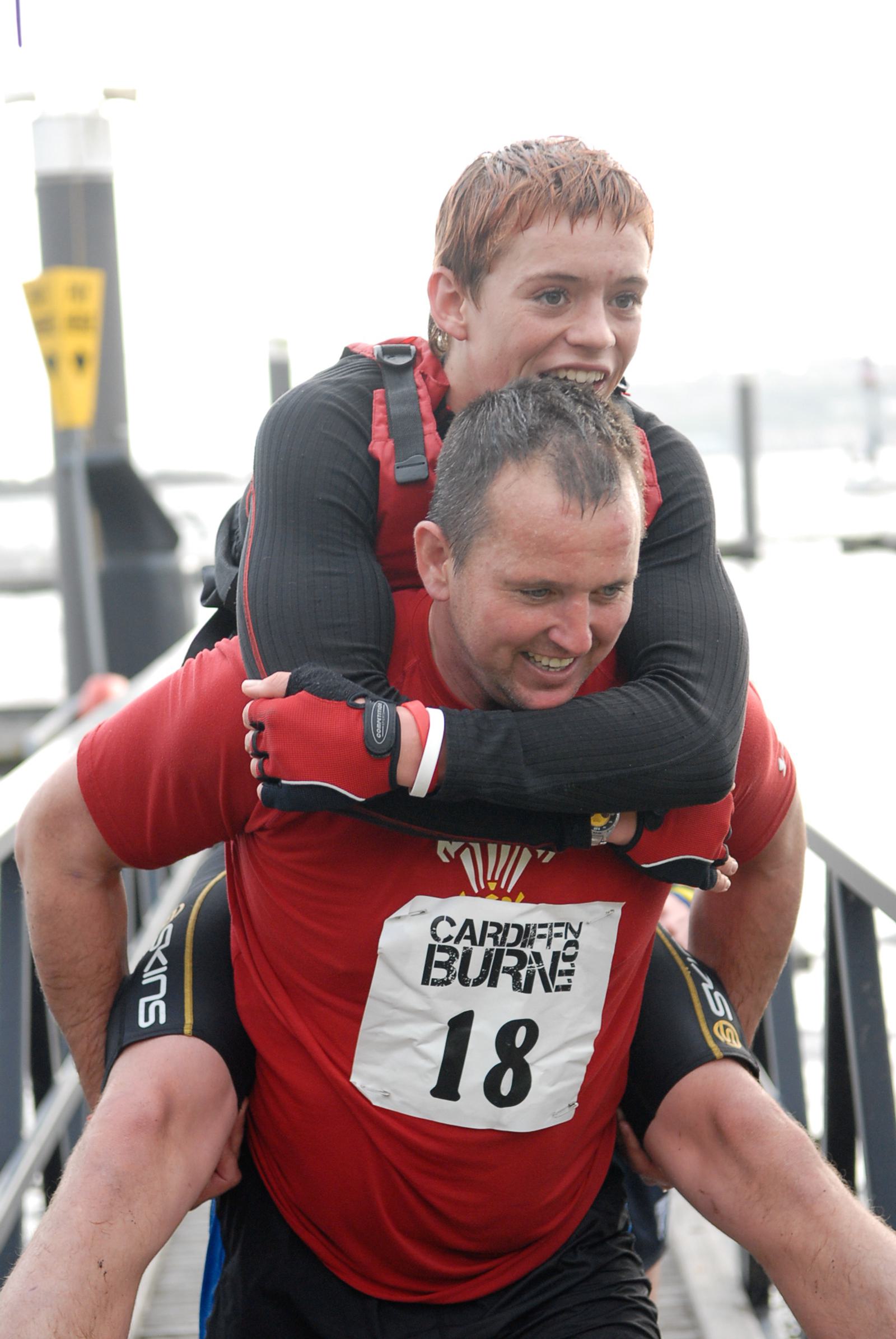 Two athletes are participating in a race. The person in the front, wearing a red shirt with race number 18, is carrying another competitor on their back. Both are smiling and appear to be enjoying the event, with a waterfront and docks visible in the background.