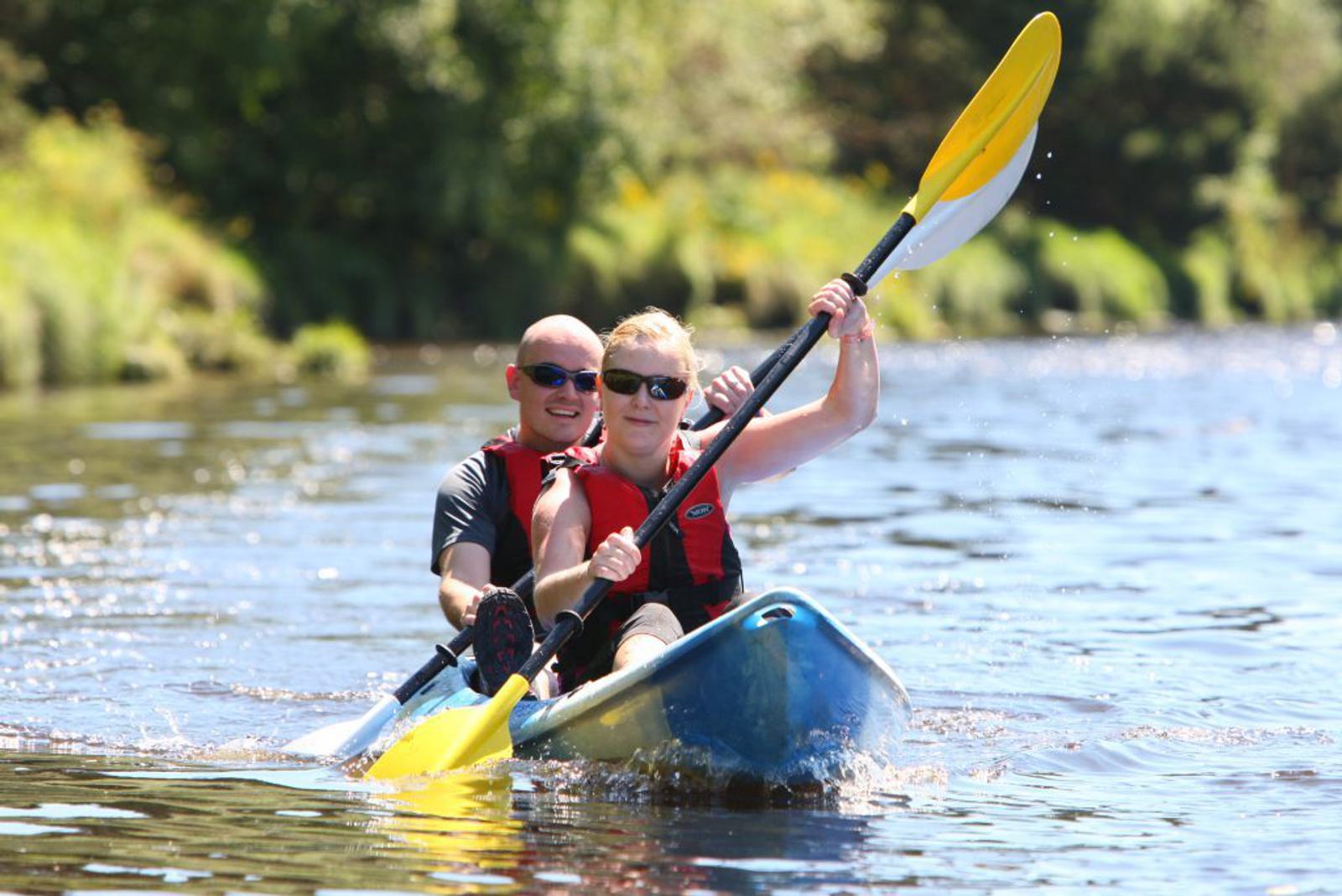 A man and a woman are kayaking together on a calm body of water. Both are wearing red life jackets and sunglasses. The woman is in the front, paddling with a yellow and white oar, while the man is behind her, holding onto his paddle. Trees are visible in the background.