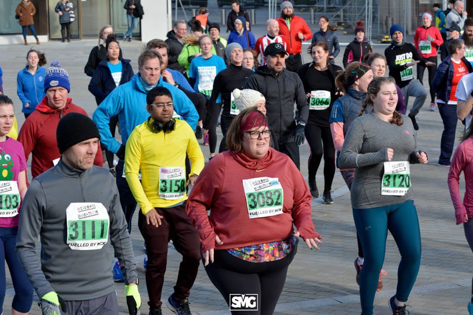 A diverse group of people running in an outdoor race. They are wearing race bibs on their shirts and layered clothing suitable for cold weather. Some participants look focused, while others appear to be enjoying the event. Background shows a cityscape with more runners.