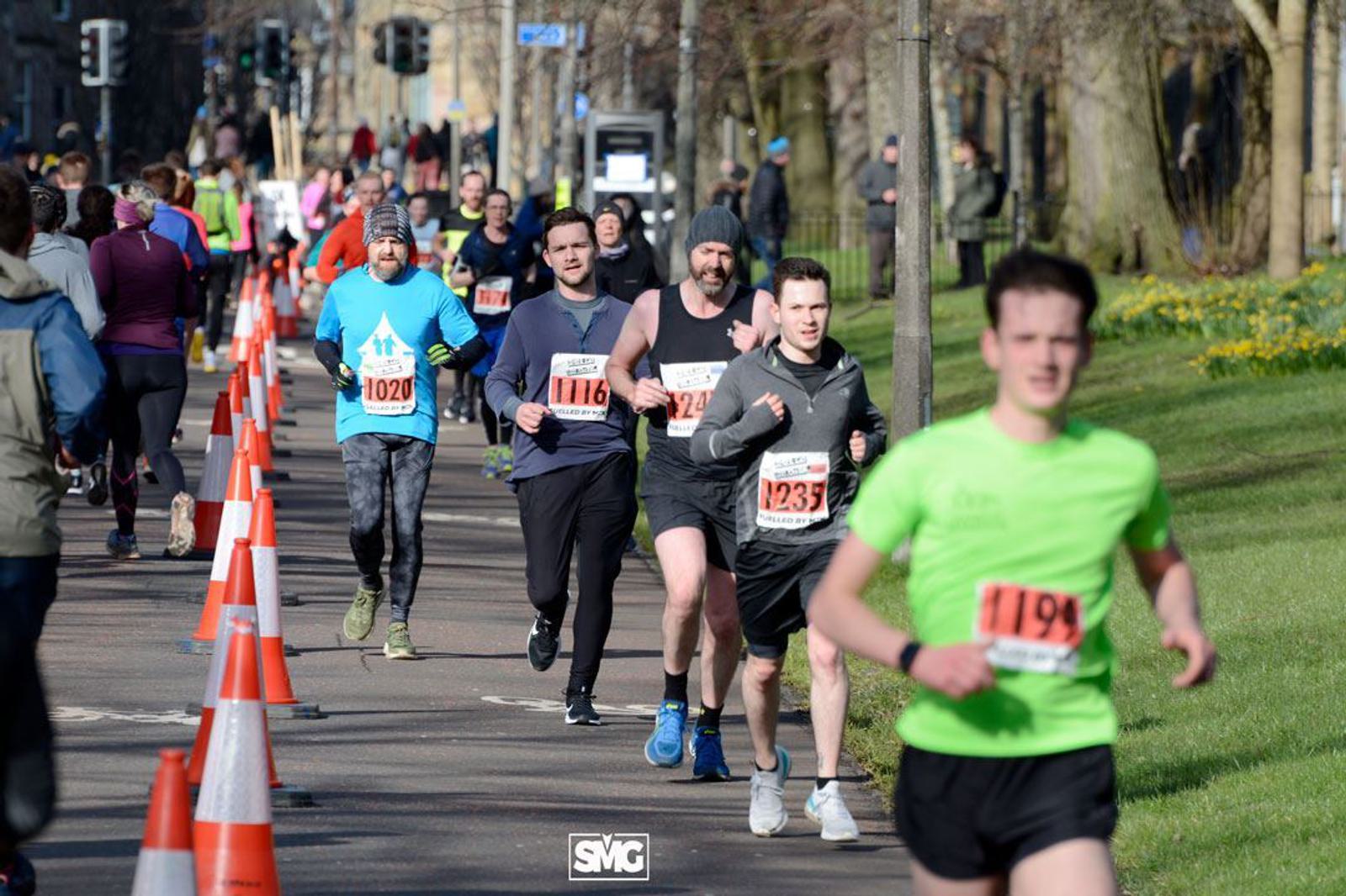 A group of runners participating in a race on a sunny day. The path is lined with orange traffic cones, and spectators are visible in the background. Some runners wear race bibs with numbers, and one runner in bright green is prominent in the foreground.