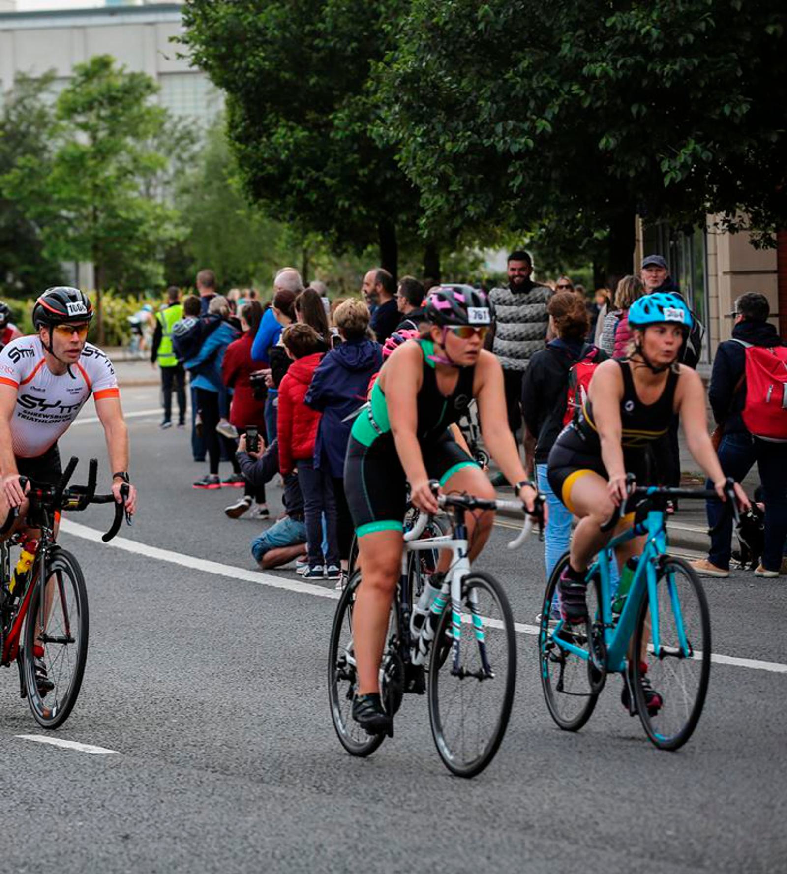 Three cyclists, wearing helmets and athletic attire, ride on a road during a race. Spectators are standing along the side of the road, watching and cheering. The background includes trees and buildings. The cyclists are focused and appear to be in motion.