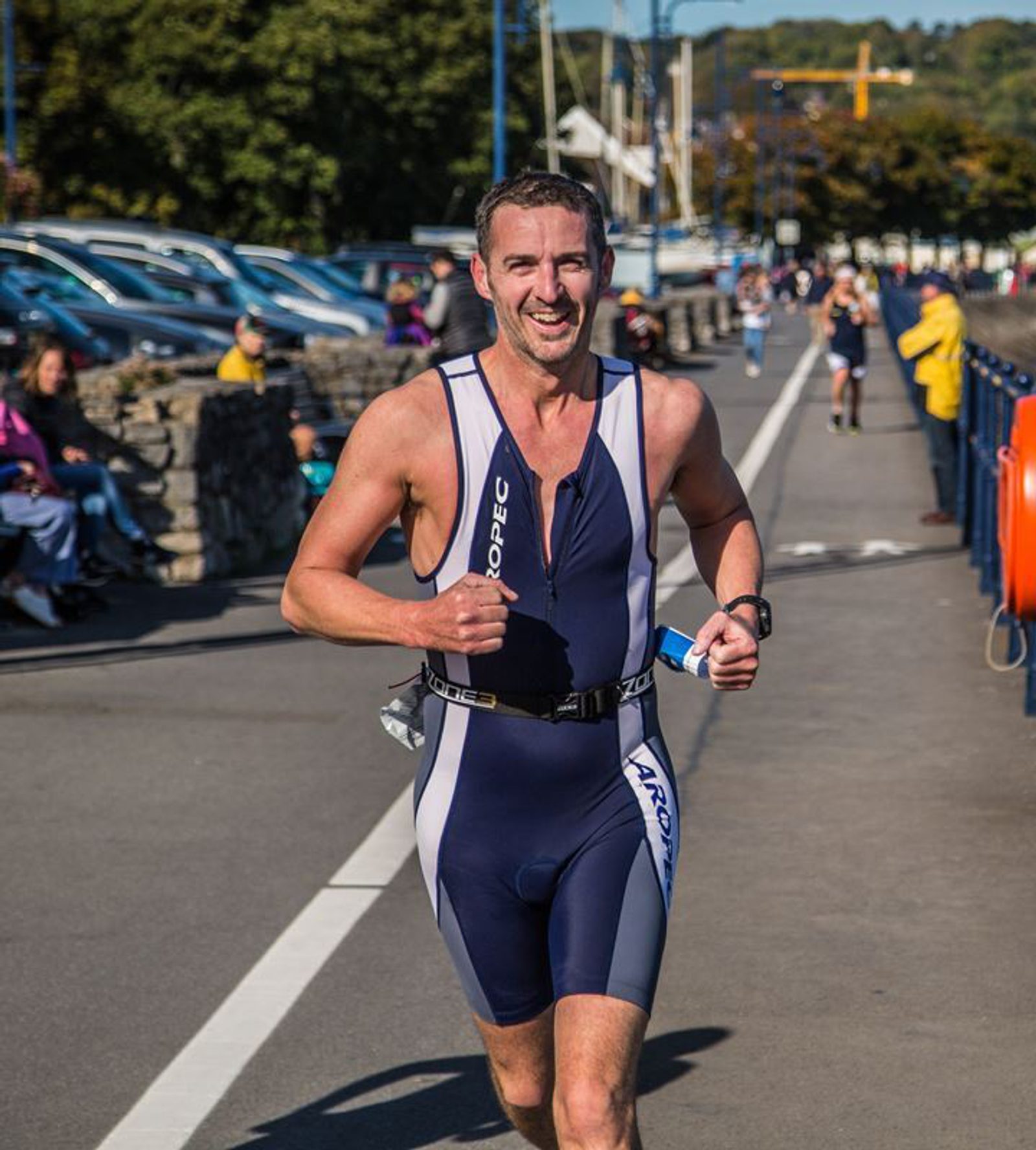 A man wearing a navy and white triathlon suit is running on a paved path during a race. He is smiling and holding a small water bottle. There are spectators seated along the side and other participants in the background. Trees and parked cars are visible.