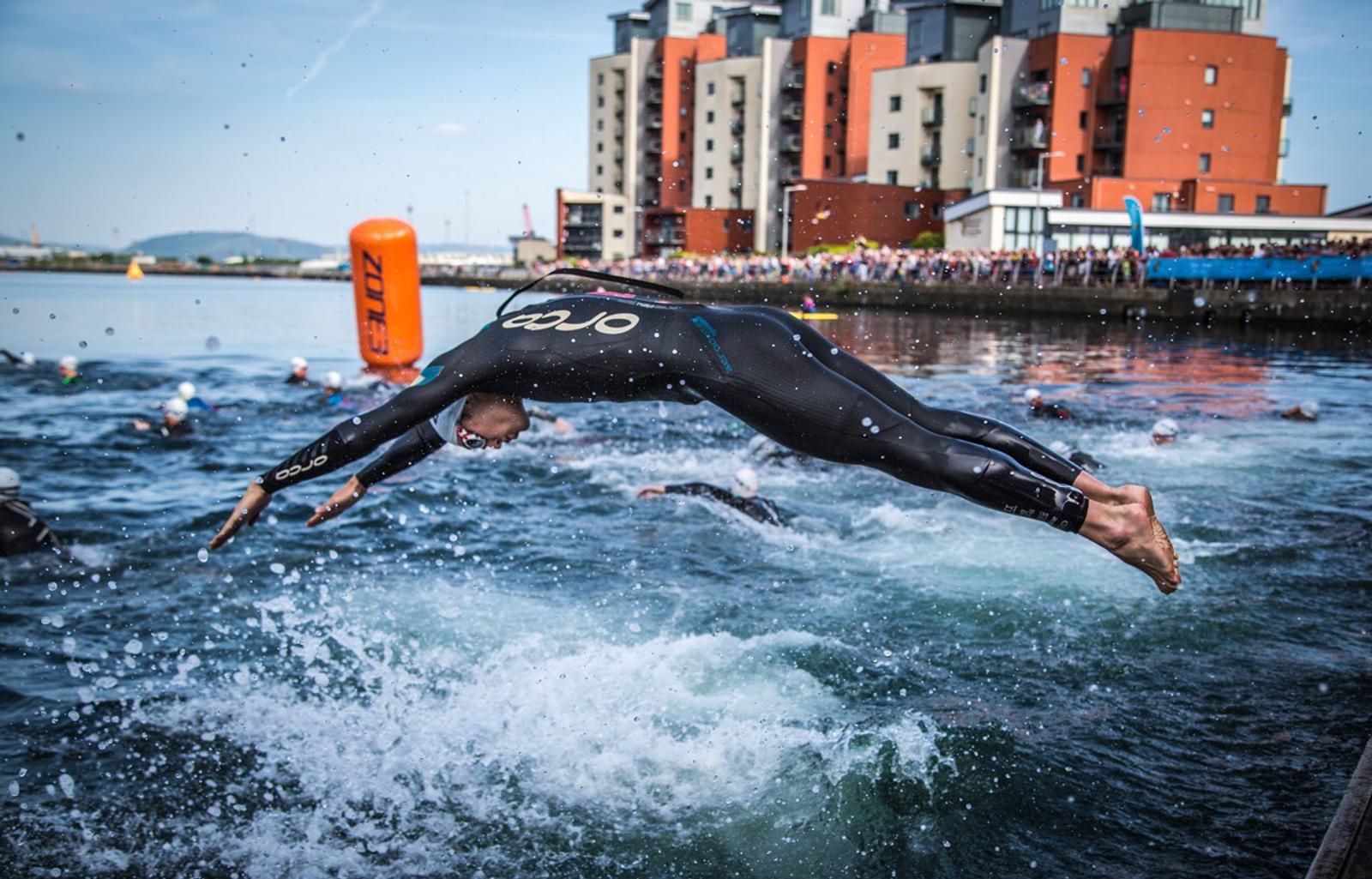 A triathlete in a black wetsuit dives into a body of water, joining a group of swimmers. The skyline features modern brick and glass buildings, and spectators line the waterfront. An orange buoy marks the swimming course.