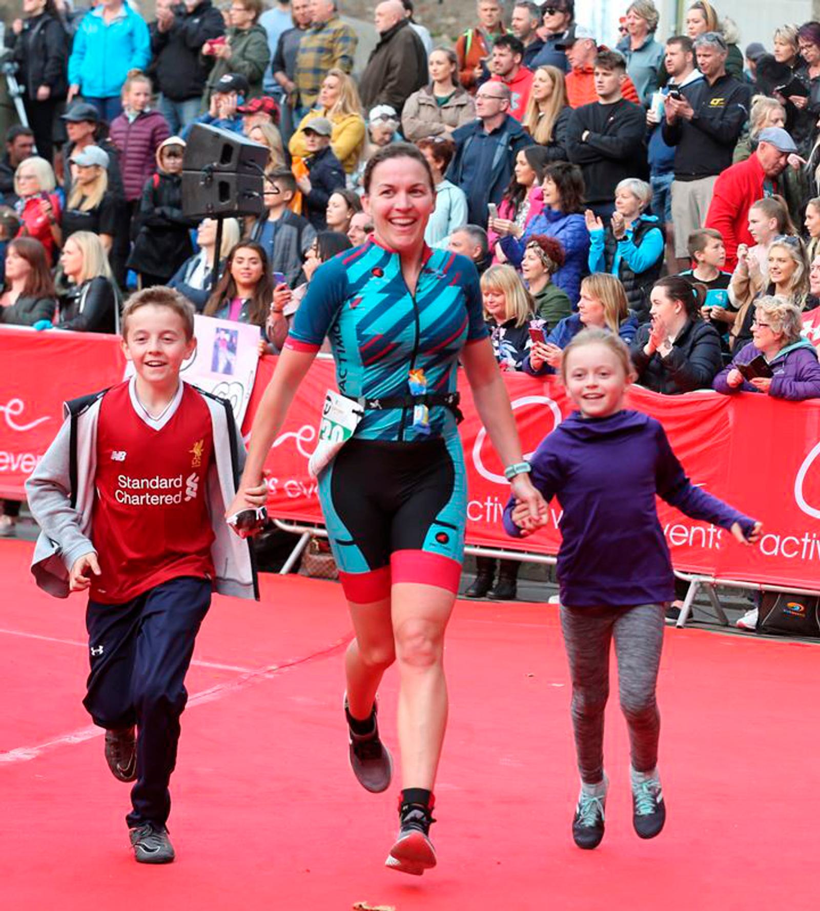A smiling woman in athletic wear crosses a finish line, holding hands with two children, all running. The boy on the left wears a Liverpool FC jersey, and the girl on the right wears a purple hoodie. A crowd of spectators and red barriers are visible in the background.