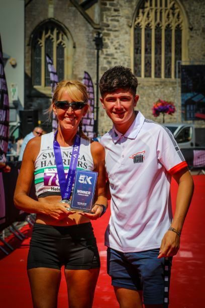 A woman in a Swansea Harriers running kit and sunglasses stands beside a younger man wearing a white shirt. She’s holding a 5K race winner’s trophy and is smiling. Both are on a red carpet, with a historic stone building in the background.