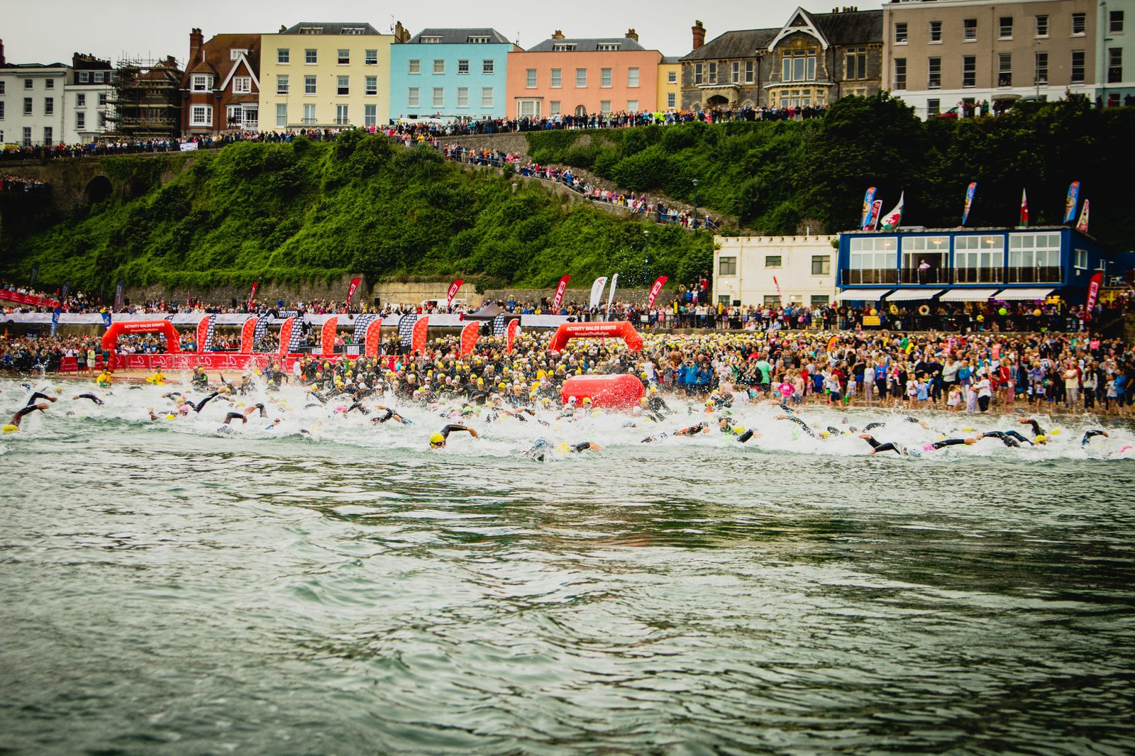 A large group of athletes begins a swimming race from a crowded beachfront. Spectators line the beach, cheering. Behind them are colorful buildings and dense greenery on the hill. Several red buoys and banners are visible in the water and onshore.