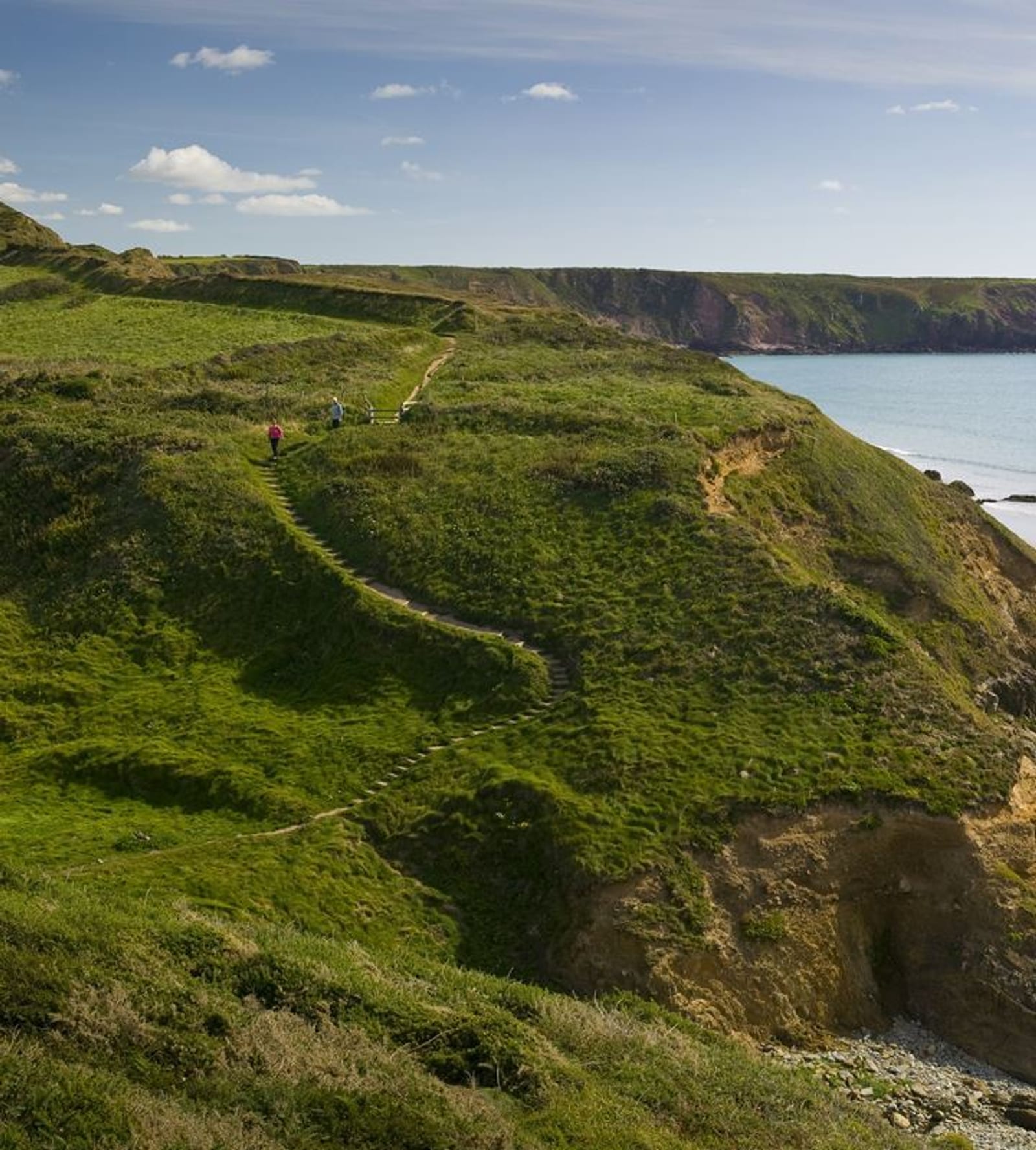 A scenic coastal landscape showing a narrow, winding path leading up a grassy hillside. Two people are walking along the path towards the top of the hill. The sea is visible on the right, with cliffs and a partly cloudy sky in the background.