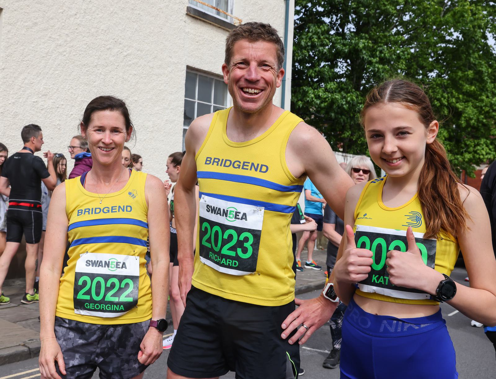 Three runners wearing yellow and blue "Bridgend" vests and green race numbers pose for a photo at an outdoor event. From left to right, their race numbers are 2022, 2023, and 2023. The girl on the right gives a thumbs-up gesture.