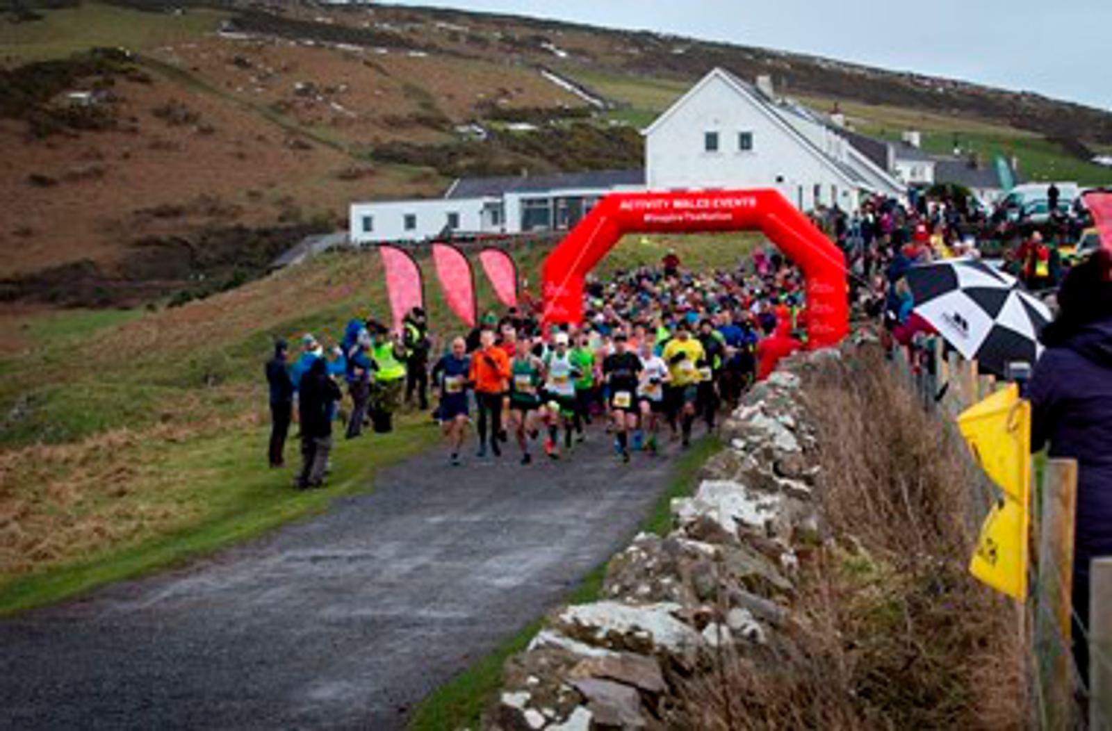A large group of runners in colorful attire start a race, passing under a red inflatable archway reading "RunWithWendy PILLAGE-RUN." Spectators with umbrellas and event staff line the path as the race begins in a rural, hilly area with hills and houses in the background.