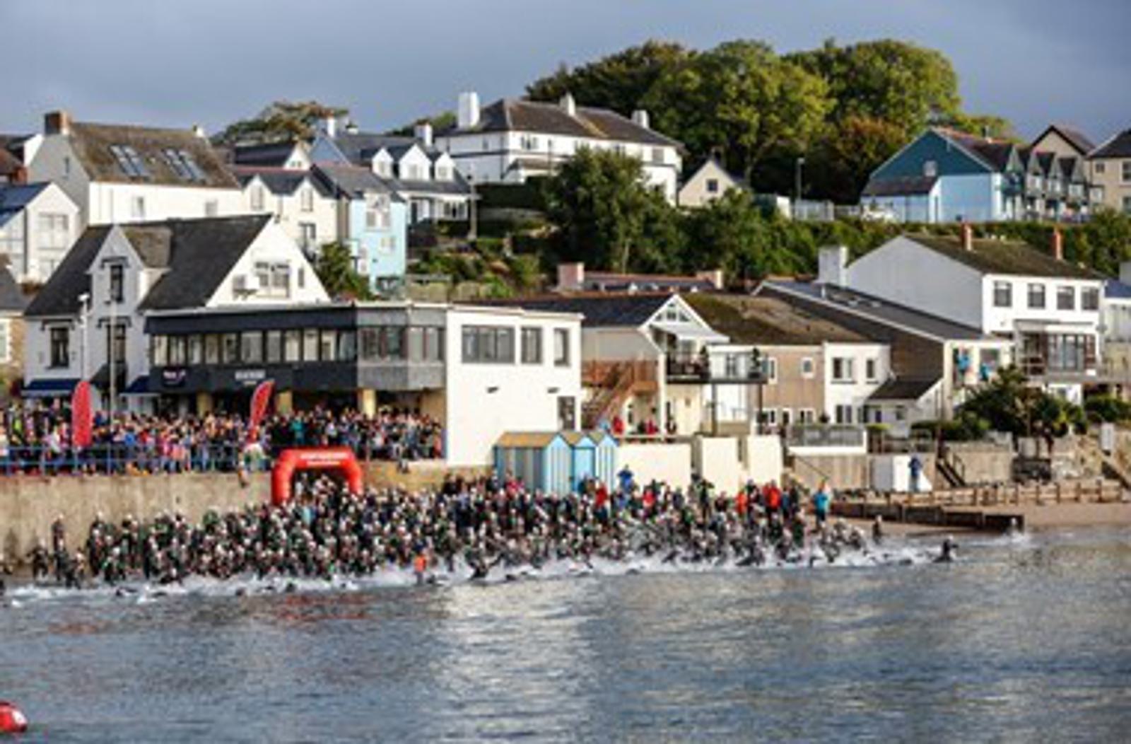 A large group of swimmers, wearing wetsuits and swimming caps, enter the water from a crowded seafront during a race. In the background, there are several houses and buildings on a hillside, with onlookers gathered along the shore to watch the event.