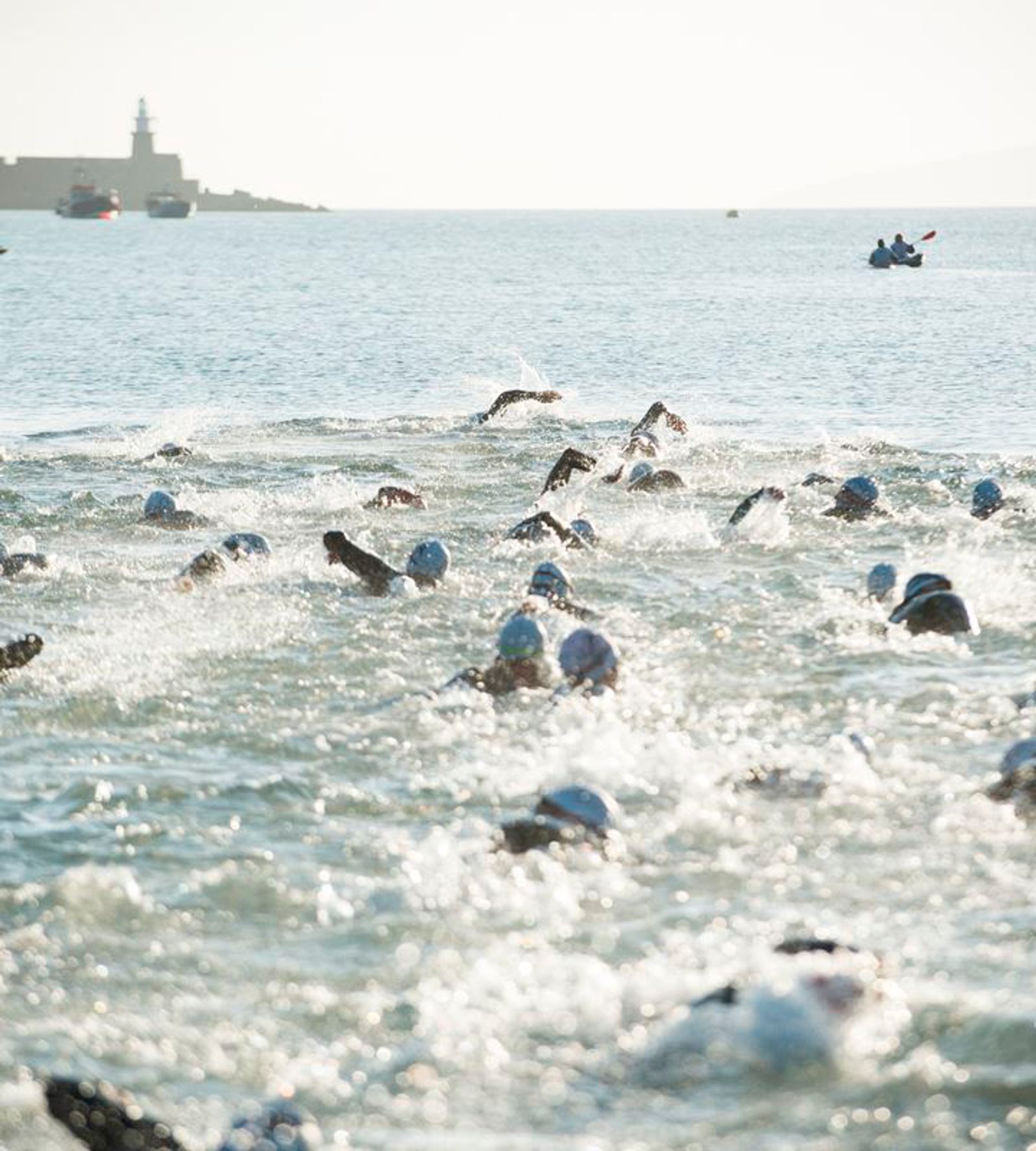 A large group of swimmers wearing swim caps and goggles competes in an open water swim, creating splashes in the ocean. The background shows a distant shoreline with a lighthouse and some people on a boat.
