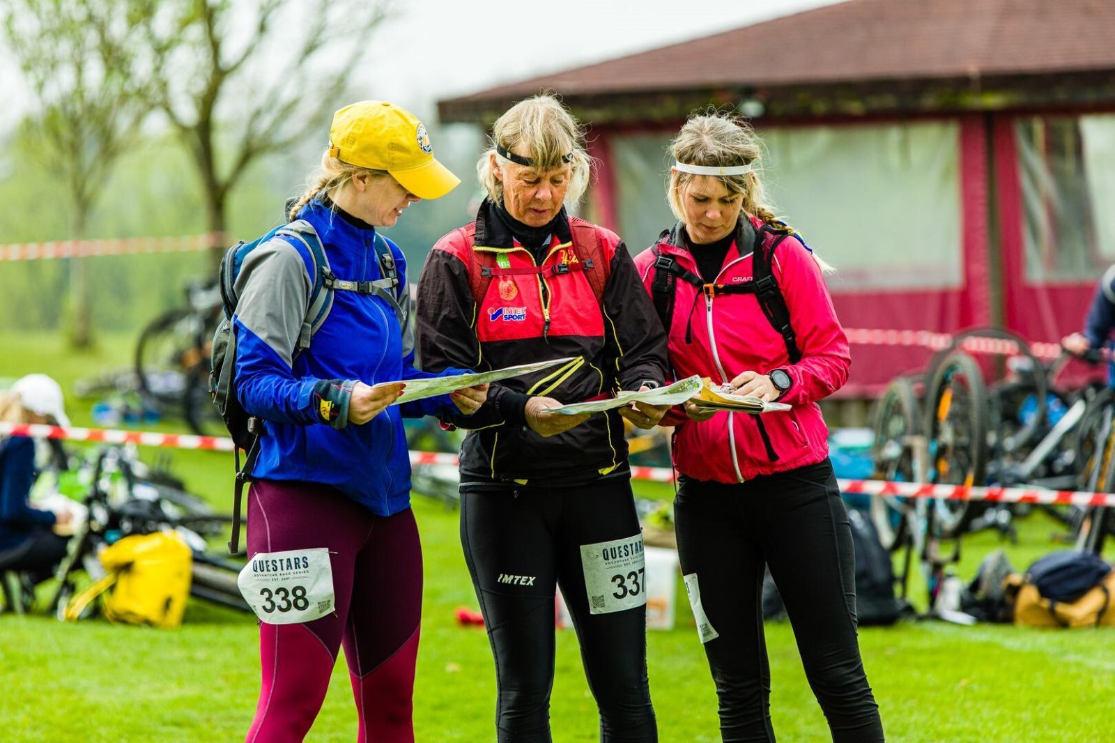 Three women in athletic gear and matching headbands stand on grass, examining maps. They wear number bibs 331, 336, and 338, indicating participation in an event. Bicycles and a red tent are in the background, suggesting a race or outdoor activity.