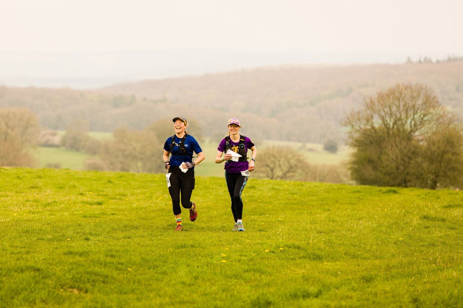 Two women in athletic gear run side-by-side on a grassy field during a daylight outdoor event. Both wear caps and race bibs, suggesting participation in a race or marathon. The background features rolling hills and trees under a hazy sky.