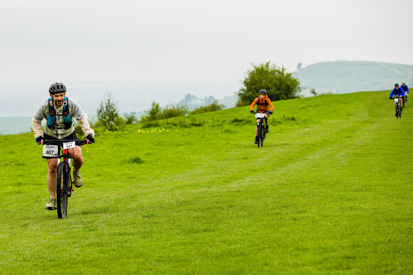 Three cyclists ride on a grassy hill during an outdoor bike race. The cyclist in the foreground wears a helmet and race number 467. Two other cyclists follow behind, with the background featuring rolling hills and a cloudy sky.