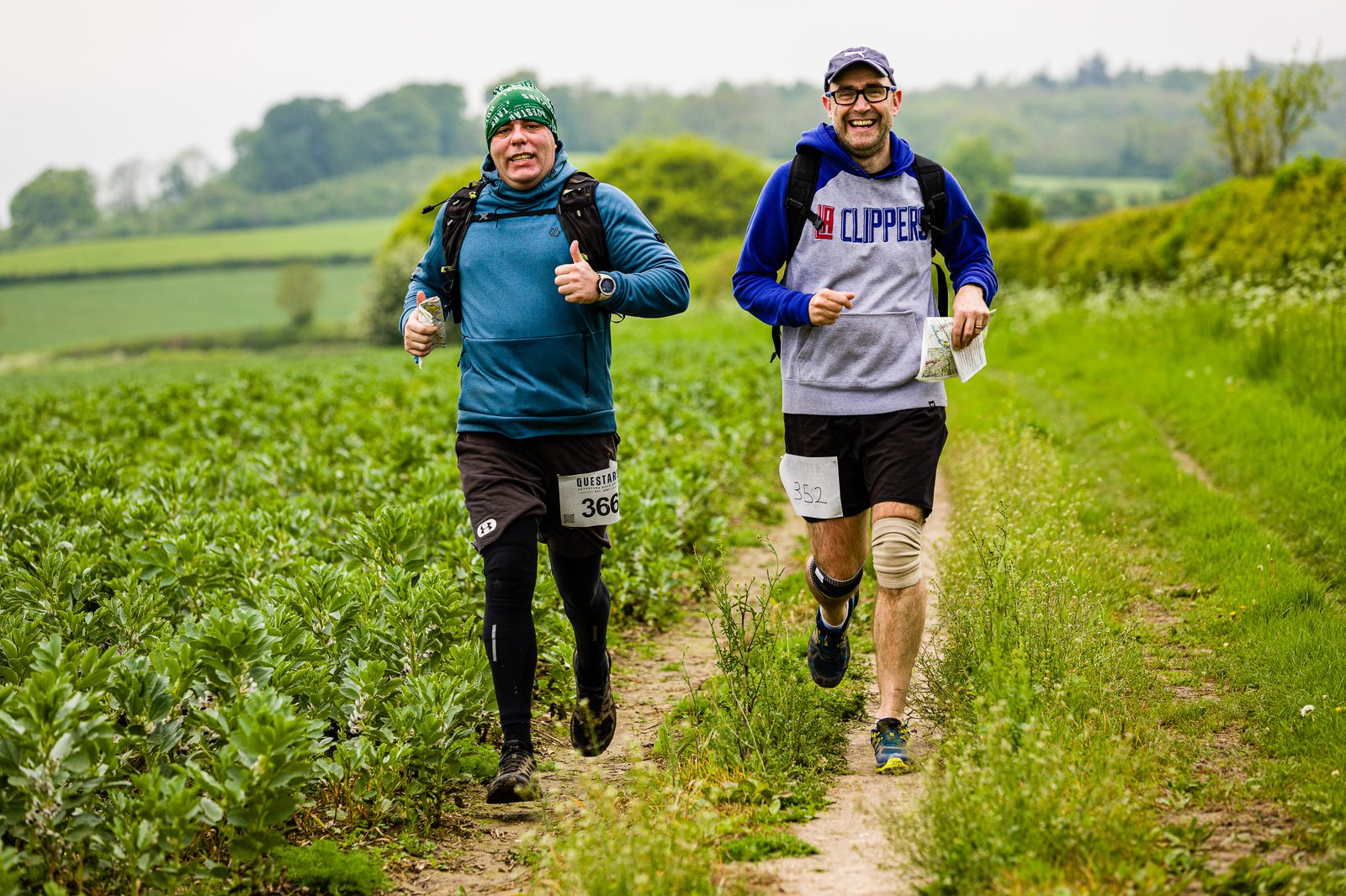 Two men are running outdoors on a trail between green fields. One is wearing a teal jacket and black pants, while the other is dressed in a blue and gray jacket and black shorts with a knee brace. Both are smiling and holding maps. Bib numbers are pinned to their clothes.