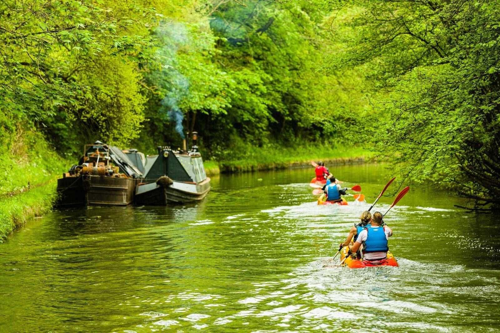 A group of kayakers in bright blue life jackets paddle along a green, lush river. Ahead, two old boats anchored at the riverbank emit smoke. The surrounding foliage is dense and vibrant, creating a serene, immersive nature scene.