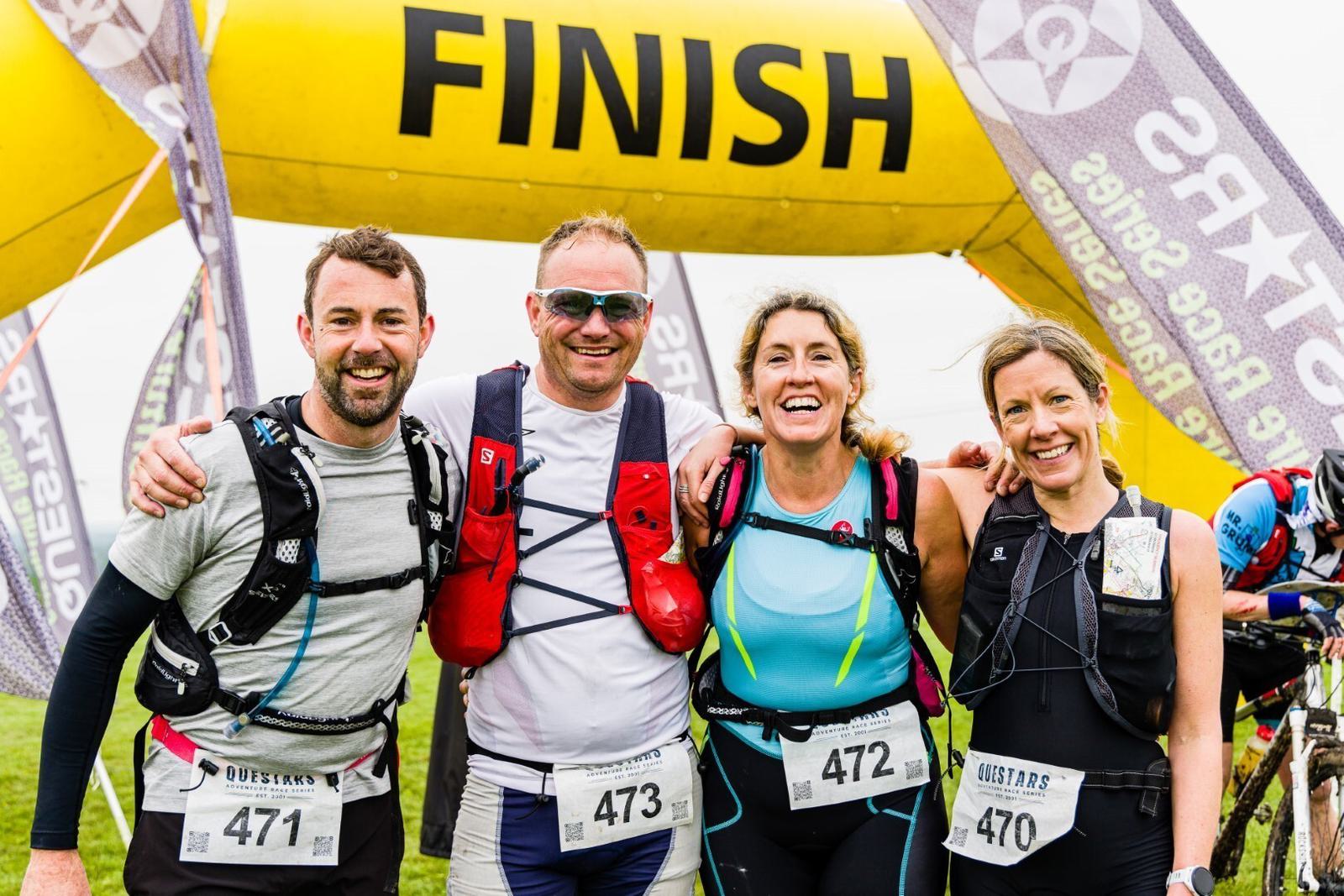 Four smiling people stand together posing for a photo under a yellow "FINISH" arch after completing a race. They wear athletic gear, race bibs with numbers 471, 473, 472, and 470, and hydration packs. More participants and a runner can be seen in the background.