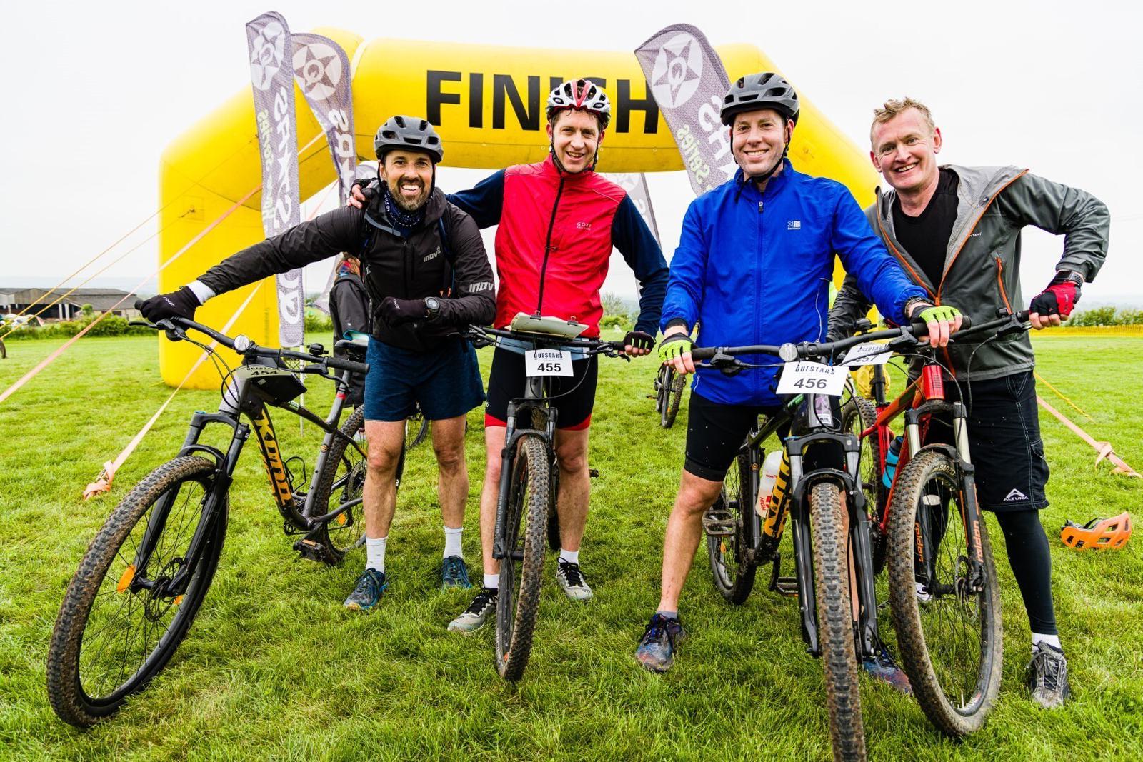 Four men with bicycles stand together, smiling at the camera near a yellow "FINISH" arch. All wear helmets and athletic clothing. One holds his bike by the handlebars; the others have their bikes positioned in front. The grass field and banners are visible in the background.