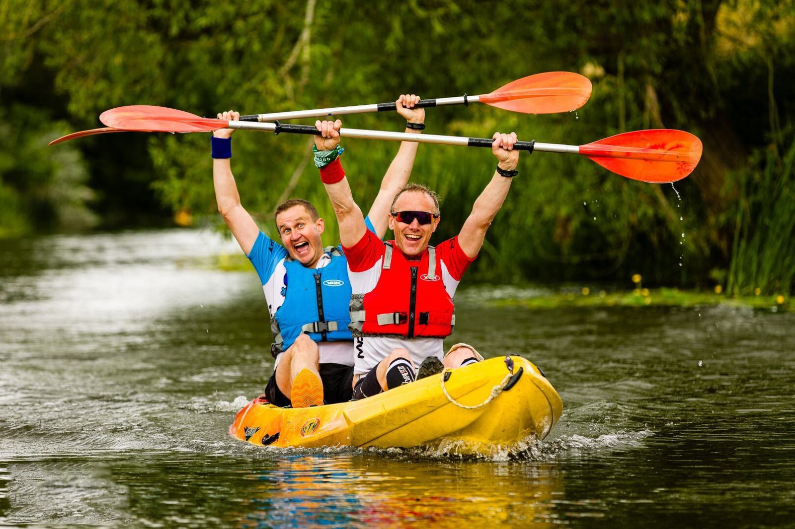 Two people in life jackets, one in blue and the other in red, paddle together in a yellow kayak on a river. Both are smiling and raising their paddles triumphantly above their heads. The surrounding area is lush with greenery.