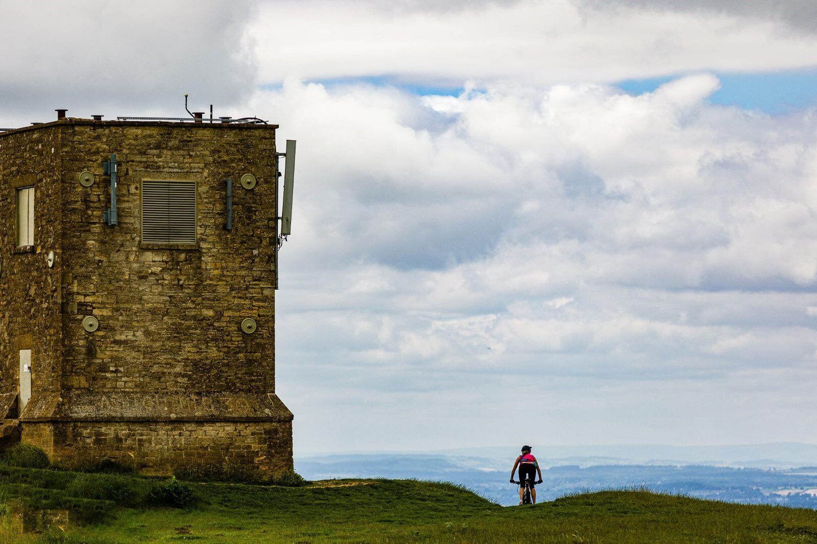 A cyclist wearing a pink jacket rides away from a large, stone building with various antennae against a backdrop of a partly cloudy sky and distant hills. The scene suggests a remote and elevated location.