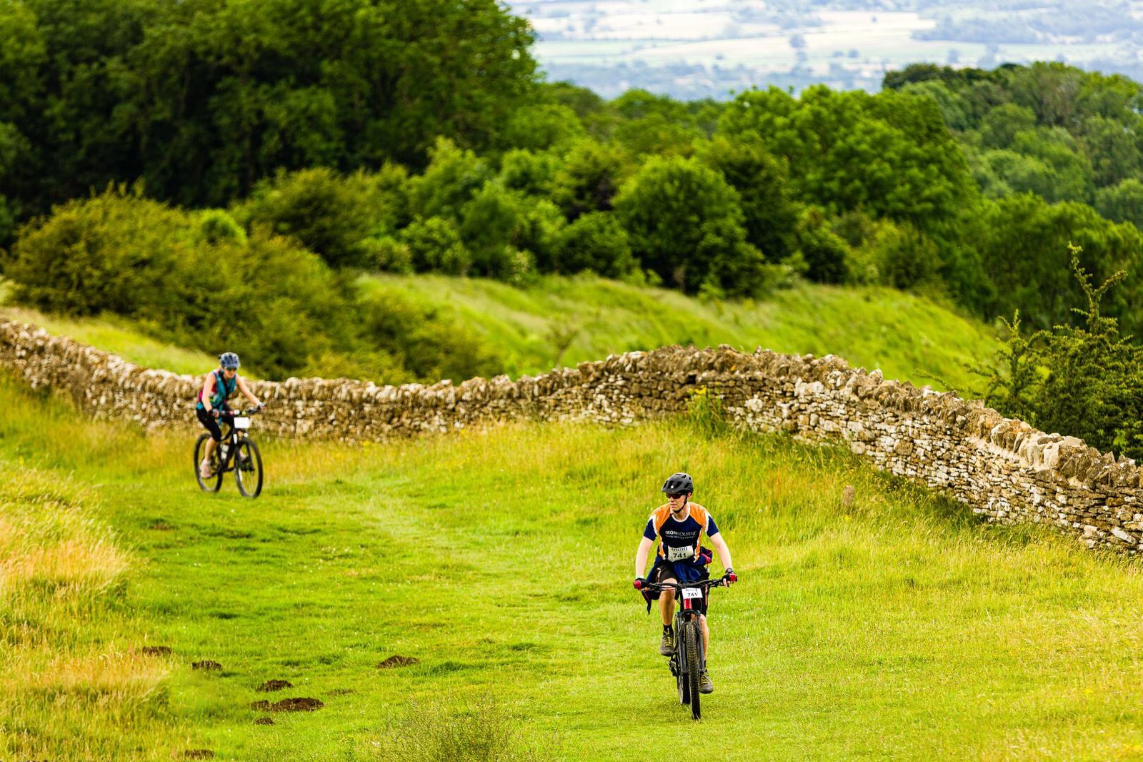 Two cyclists ride on a grassy trail beside a stone wall, with lush green fields and trees in the background. One cyclist leads while the other follows at a distance. The scene has a vibrant, natural setting with rolling hills and a partially cloudy sky.