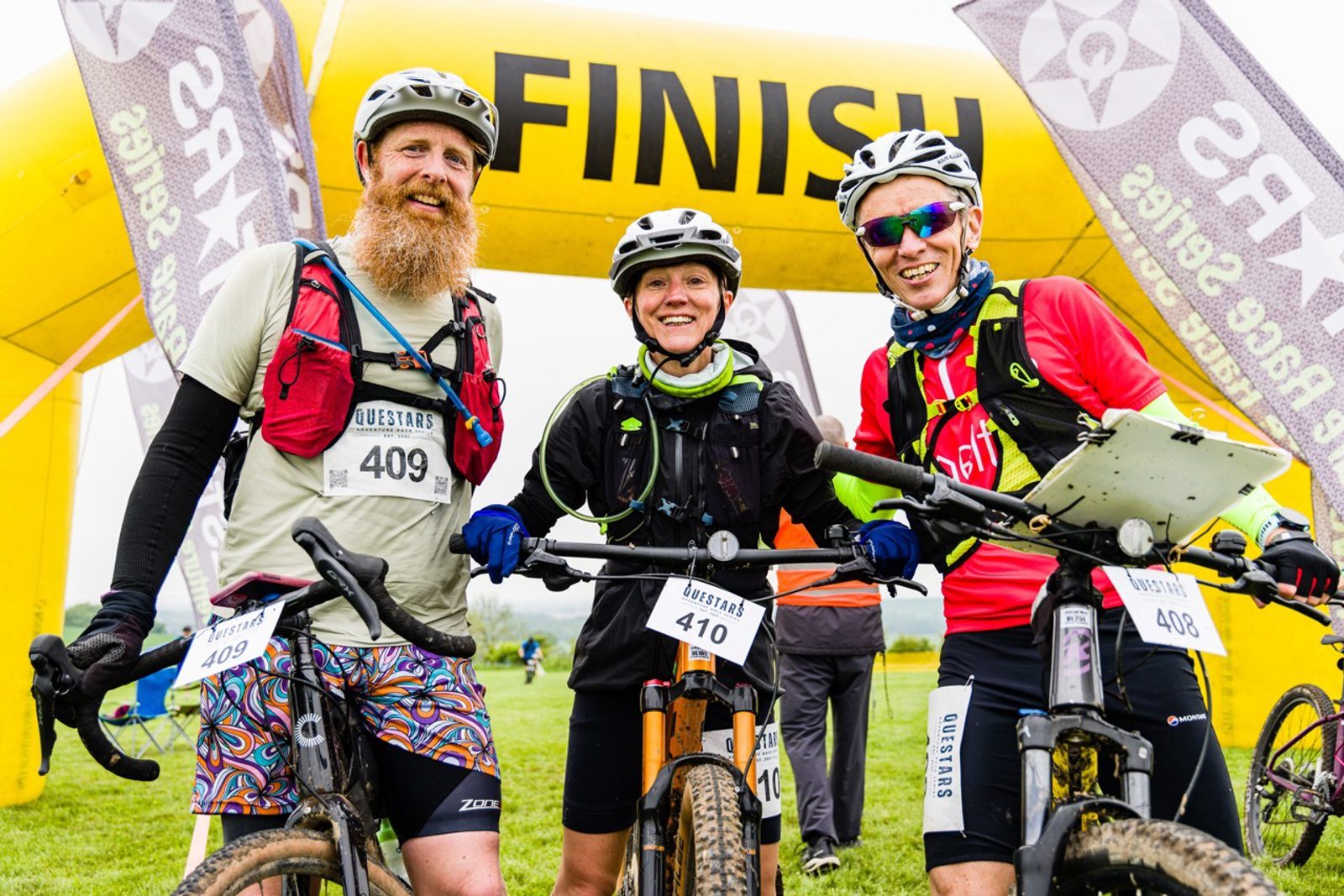 Three cyclists stand with their bikes and smile at the finish line of a race. They are wearing colorful cycling gear, race bibs (409, 410, 408), and helmets. A yellow inflatable arch with the word "FINISH" is visible behind them.