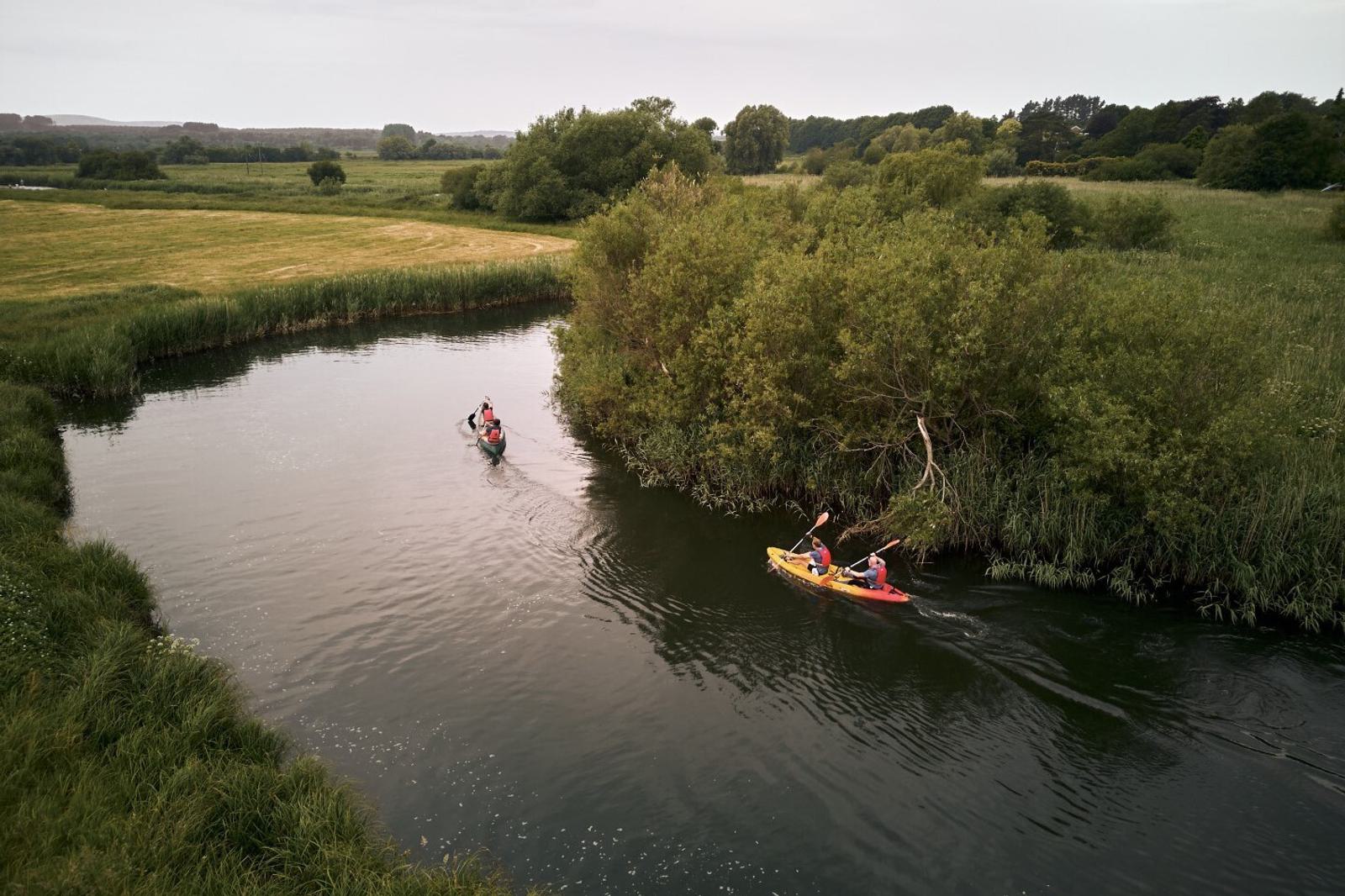 Two groups of people are kayaking on a narrow river with lush greenery on both sides. The leading group has two people in a yellow kayak, and the trailing group has two people in a red kayak. The landscape is serene, with trees and grass fields in the background.