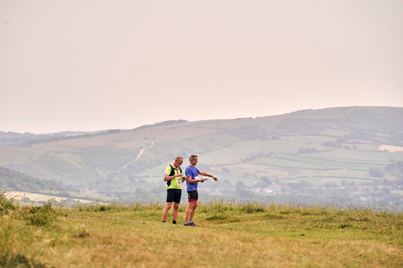 Two people are standing on a grassy hilltop, looking at a map. They are dressed in sporty clothing suitable for hiking. The background features rolling green hills under a slightly overcast sky. The scene conveys outdoor exploration and navigation.