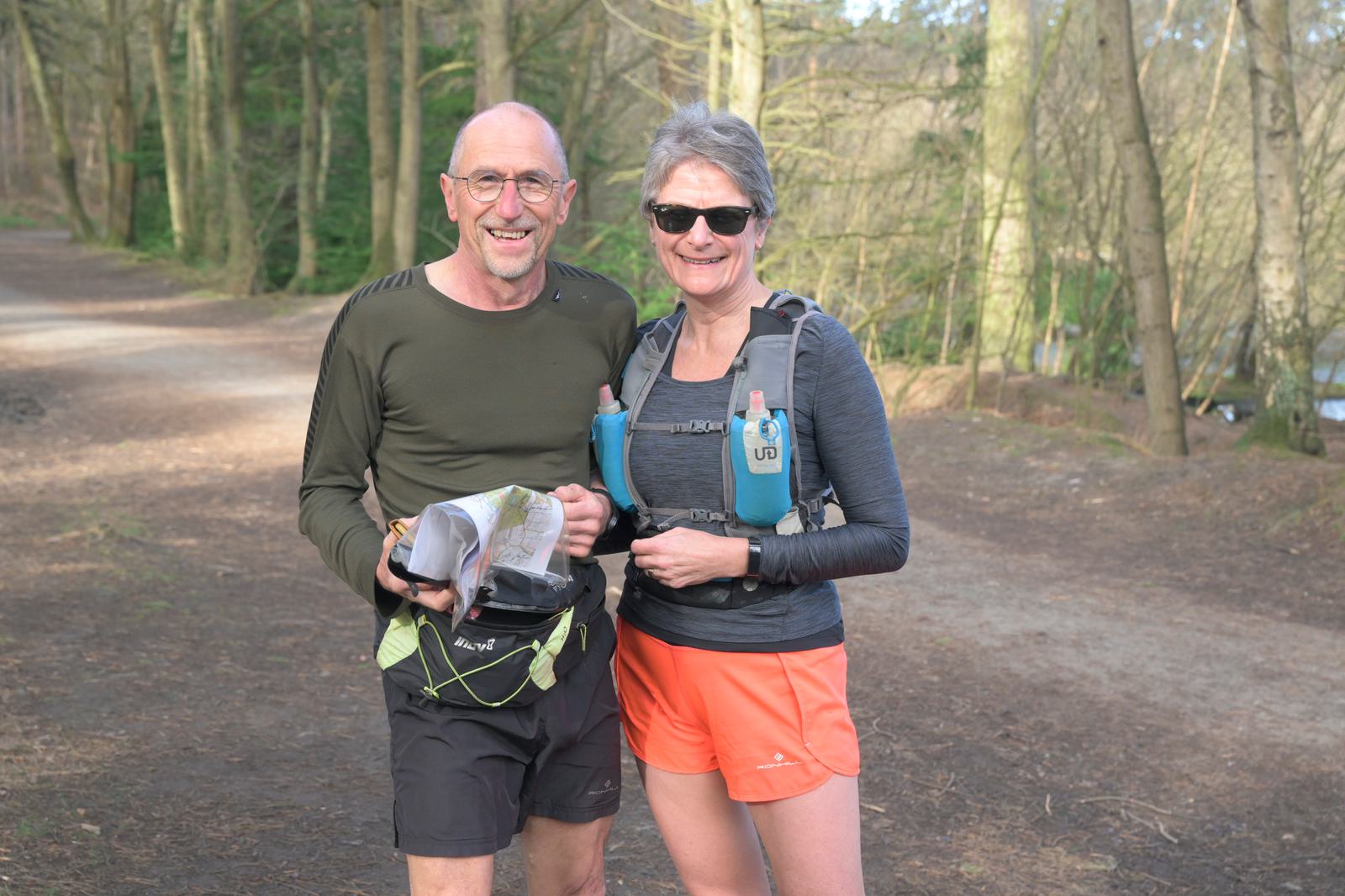 Two people in athletic gear stand smiling on a forested path. One is holding a map and wearing a green long-sleeve shirt and black shorts, while the other wears a grey long-sleeve shirt and orange shorts, both with hydration packs. Trees and trail are in the background.