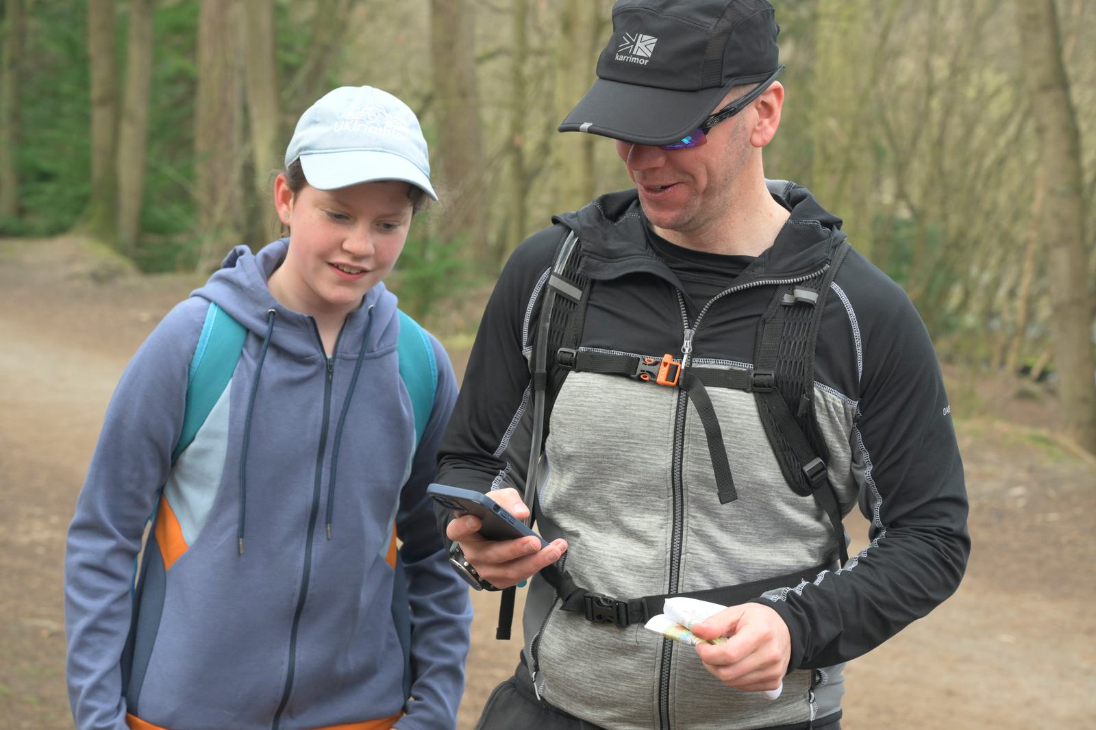 A man and a boy in outdoor gear are walking on a forest trail. The man, wearing a cap and sunglasses, is holding a smartphone and a snack, while the boy, wearing a cap and hoodie, looks on. Trees and a dirt path are visible in the background.