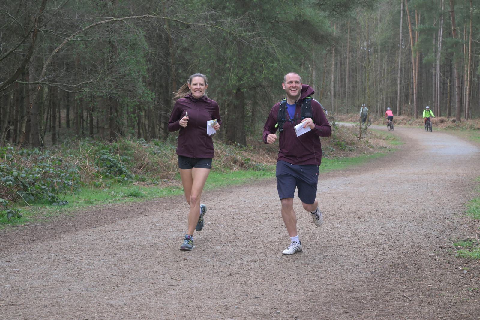 Two people are jogging on a forest trail, both smiling and holding maps. The trail is surrounded by trees and bushes. In the background, there are a few other people cycling and walking. The weather appears overcast, and the scene is peaceful and natural.