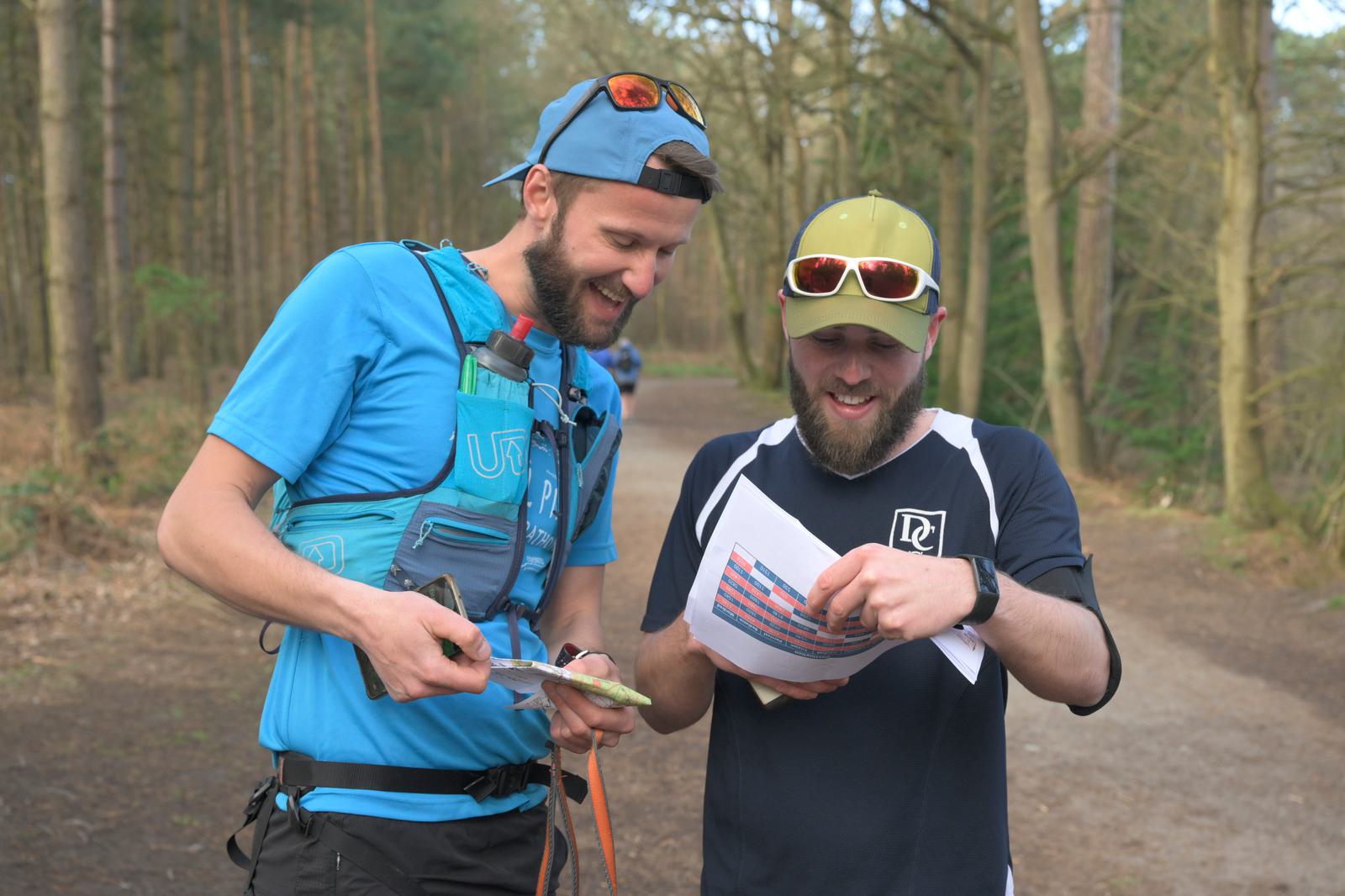 Two men in outdoor attire stand on a forest trail. Both wear caps and sunglasses. The man on the left, dressed in blue, holds a map and a compass, while the man on the right, dressed in black, points to a map or paper and smiles. Sunlight filters through the trees.